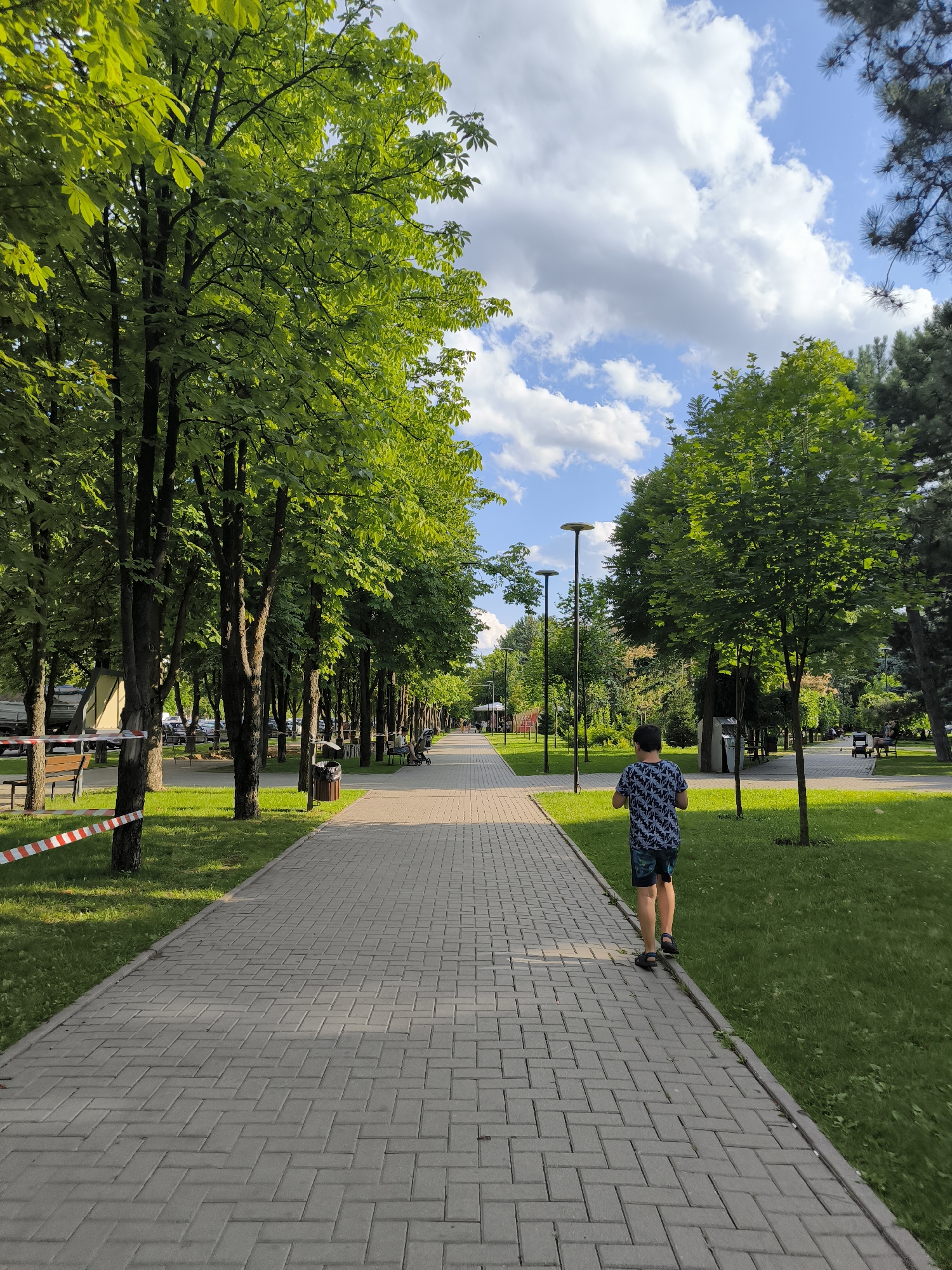 This image shows a paved walkway lined with trees in a park. A young boy wearing a patterned shirt and shorts is walking away from the camera on the right side of the path. To the left of the path, there is a grass area with benches, a trash can, and red and white caution tape tied around a tree. In the background, more trees, streetlights, and a few people are visible. The sky is blue with scattered white clouds. This scene is consistent with areas in parks located in Chișinău, Moldova.
