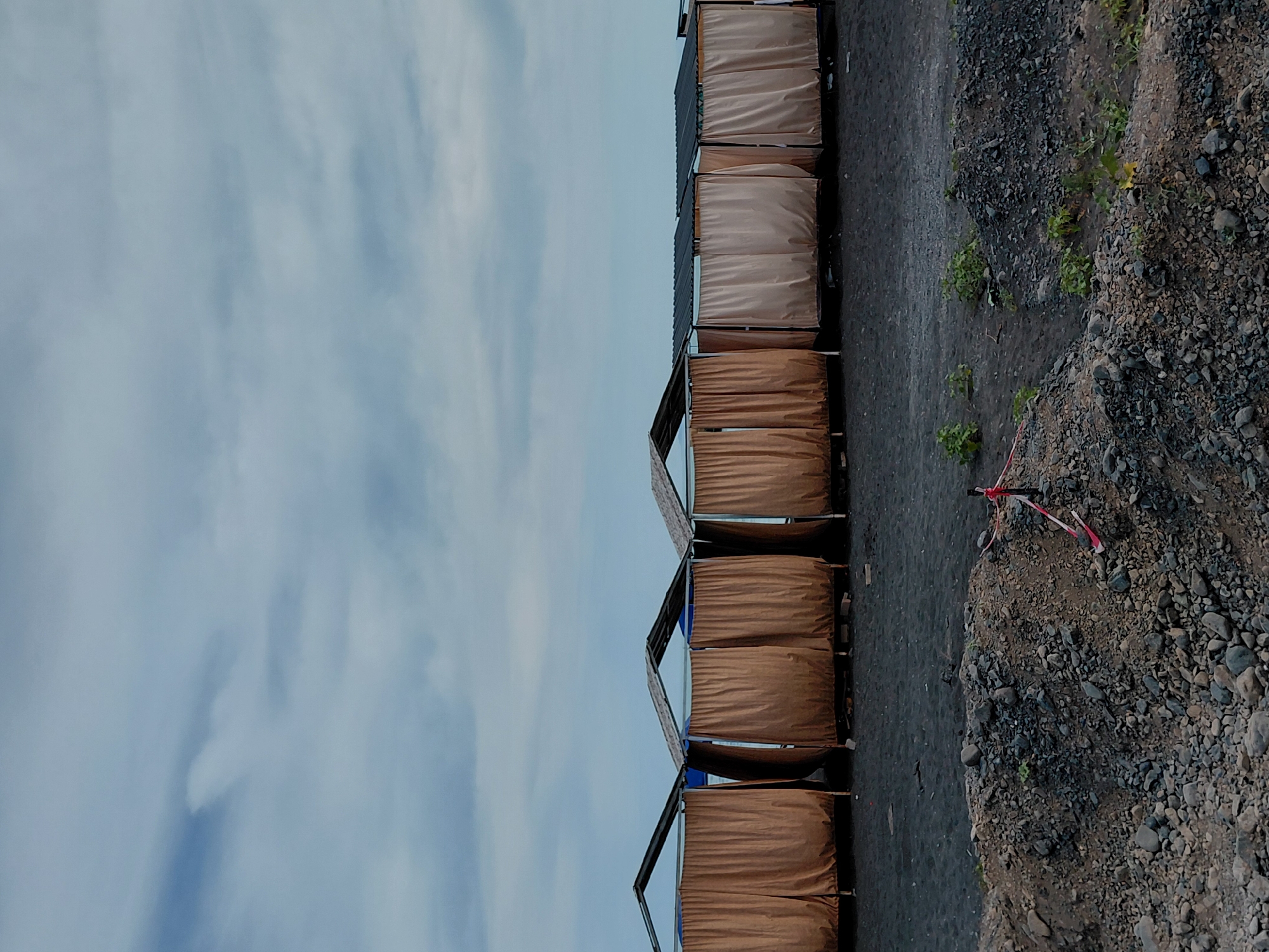 The image depicts a row of closed beach cabanas with brown and beige curtains, set against a backdrop of a cloudy sky and a dark sandy beach. The cabanas have peaked roofs and appear to be constructed from metal frames. In the foreground, a pile of rocks and gravel is visible, with some sparse green vegetation growing amongst them. A red and white striped rope or barrier is tied to a pole in the rock pile. The scene is devoid of people. The location is specified as the city of Ақши, Kazakhstan.