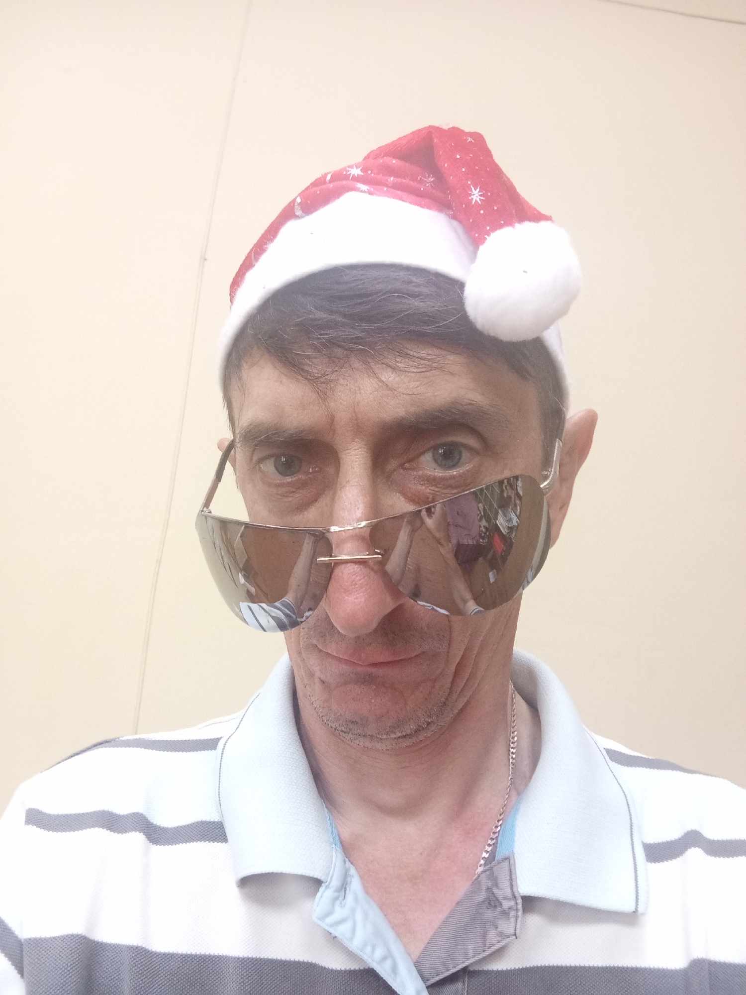A man wearing a Santa hat and sunglasses is pictured from the chest up, looking directly at the camera. The background is a plain, light-colored wall, possibly in a room. The man has brown hair, visible stubble, and is wearing a horizontally striped polo shirt with gray and white bands. The Santa hat is red with white trim and a white pom-pom, decorated with small white snowflakes. The sunglasses are large and mirrored, reflecting a blurry image of what appears to be a patterned shirt or garment. A thin silver chain is visible around his neck, tucked into his shirt collar. The image does not provide specific information that would allow for the determination of its location in Крыўлянскі сельскі Савет, Belarus.