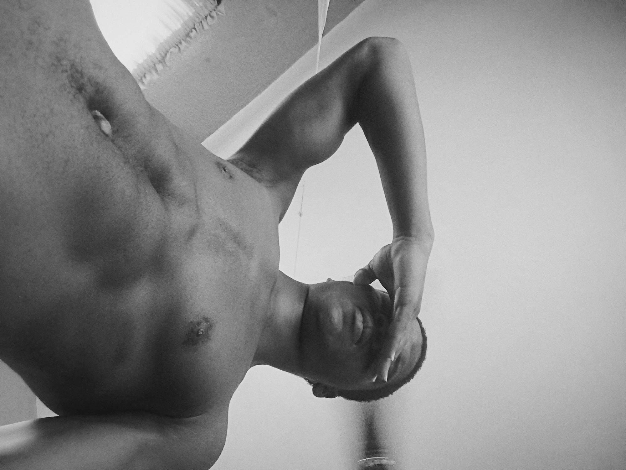 A black and white, low-angle photograph depicts a shirtless man with dark skin. His head is tilted downward, and he appears to be holding his head with his right hand. The visible portion of his torso shows some muscular definition. The background is a plain white wall. The environment and contents do not provide sufficient information to determine location in Nkpor, Nigeria.