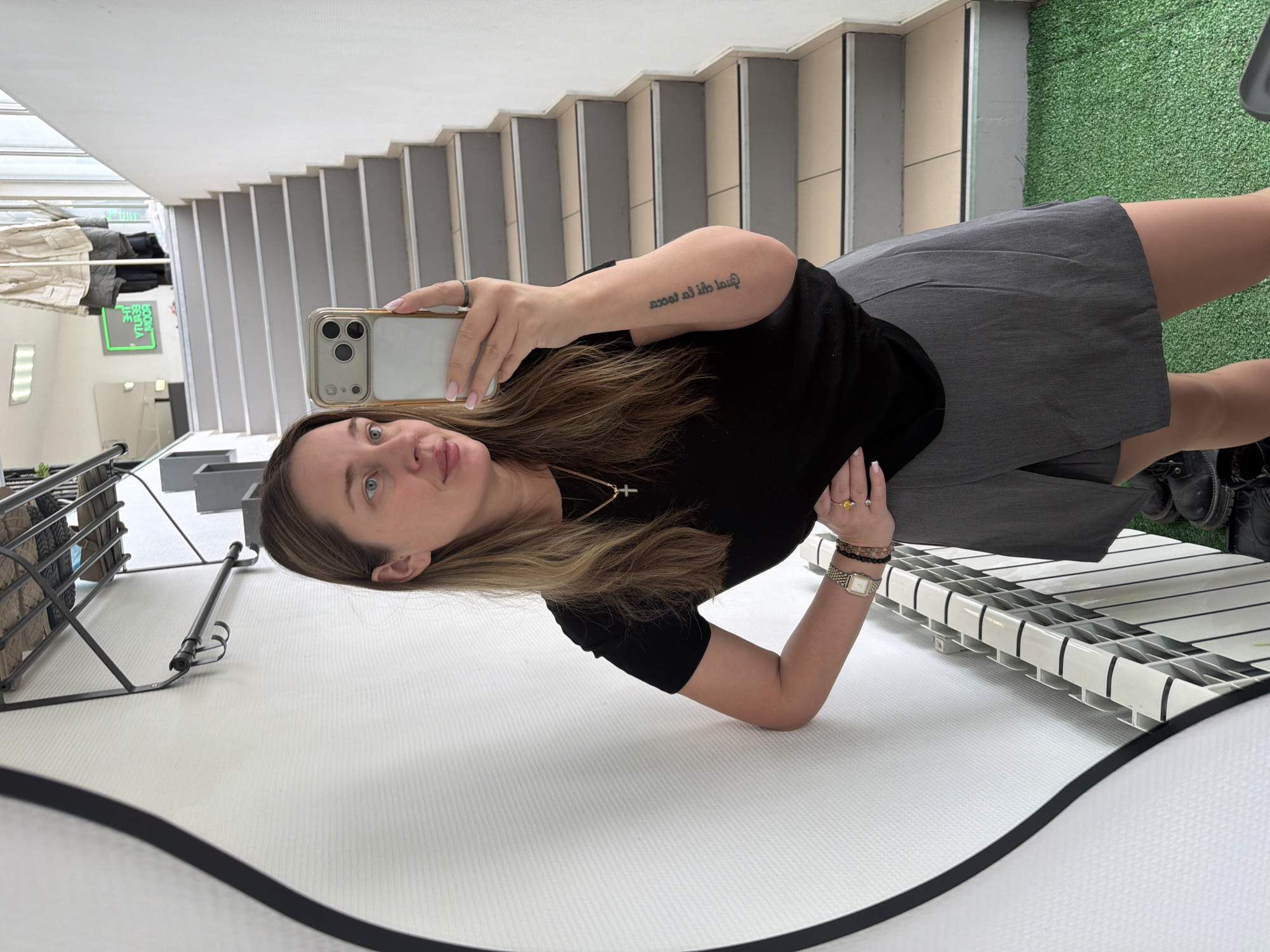 A woman is taking a selfie in a mirror, inside a building. She is wearing a black shirt, a gray skirt, and black boots. She has a tattoo on her arm, a watch, and bracelets. The background includes stairs, artificial grass, and some furniture. The location is not discernible as Chaikovsky, Russia, based on the image.