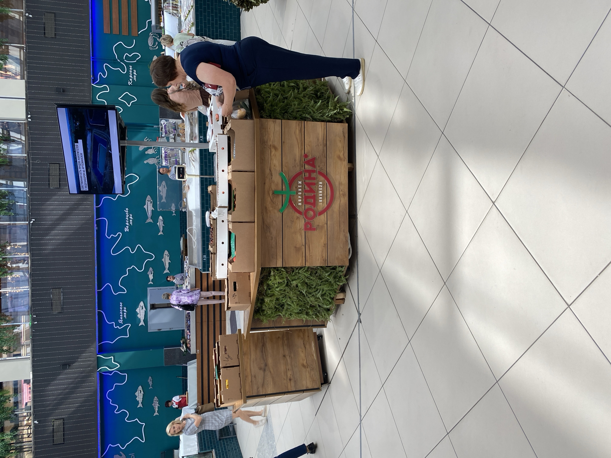 The image captures a brightly lit interior scene, likely a market or food hall, with a noticeable focus on produce and fish. A prominent green wall with white line art depicting fish and waves serves as a backdrop. In the foreground, a wooden display stand is laden with cardboard boxes filled with what appear to be fruits and vegetables. A large red circular logo with the text "ВОРОНЕЖ РОДИНА" and "ORONEZH" is visible on the front of the stand, identifying the brand as Rodina from Voronezh.
Several people are present in the scene. A woman in a checkered dress is standing to the left, looking away from the camera. A person in a purple top and light-colored pants is in the middle ground, facing a counter. To the right, a woman in a dark blue outfit is interacting with the display stand, appearing to select items. Another person, partially visible, stands behind her. In the background, various stalls and displays are visible, including one with fish on ice. A large television screen mounted on a stand displays news or informational content. The floor is tiled with a light-colored, geometric pattern. The overall atmosphere suggests a bustling commercial environment. While the prompt specifies Novaya Usman', Russia, this specific location cannot be definitively confirmed from the visual information alone.