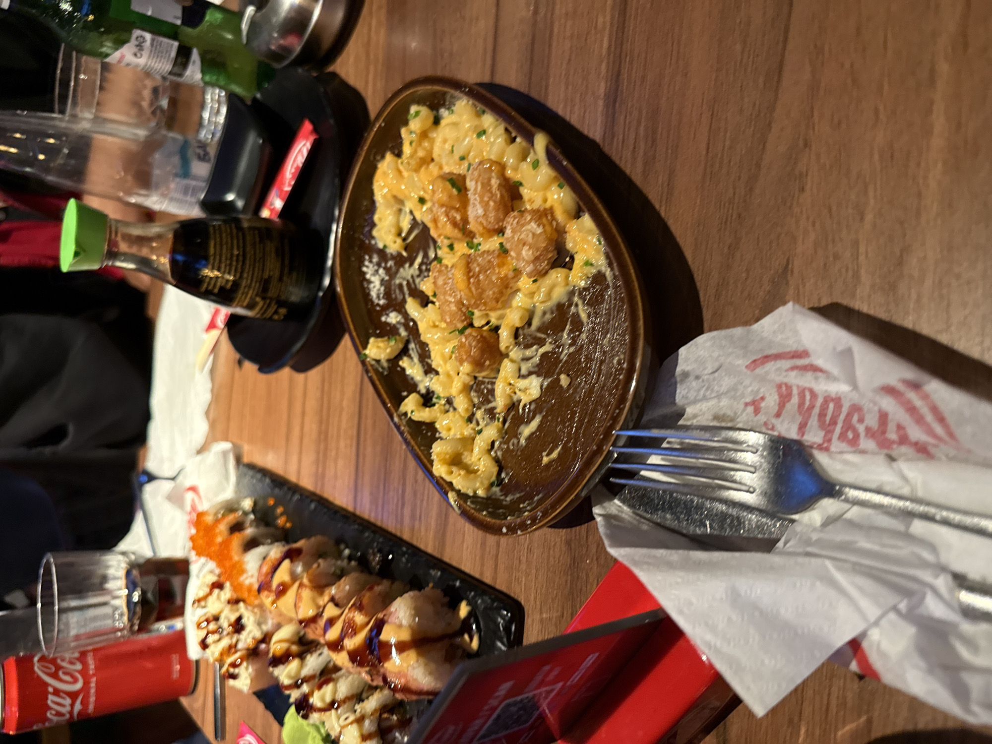 The image shows a table setting at a restaurant. A plate of macaroni and cheese with breaded toppings is present, along with a plate of sushi. There is also a soy sauce bottle, drinks, and cutlery with napkins. The location is in Burgas, Bulgaria.
