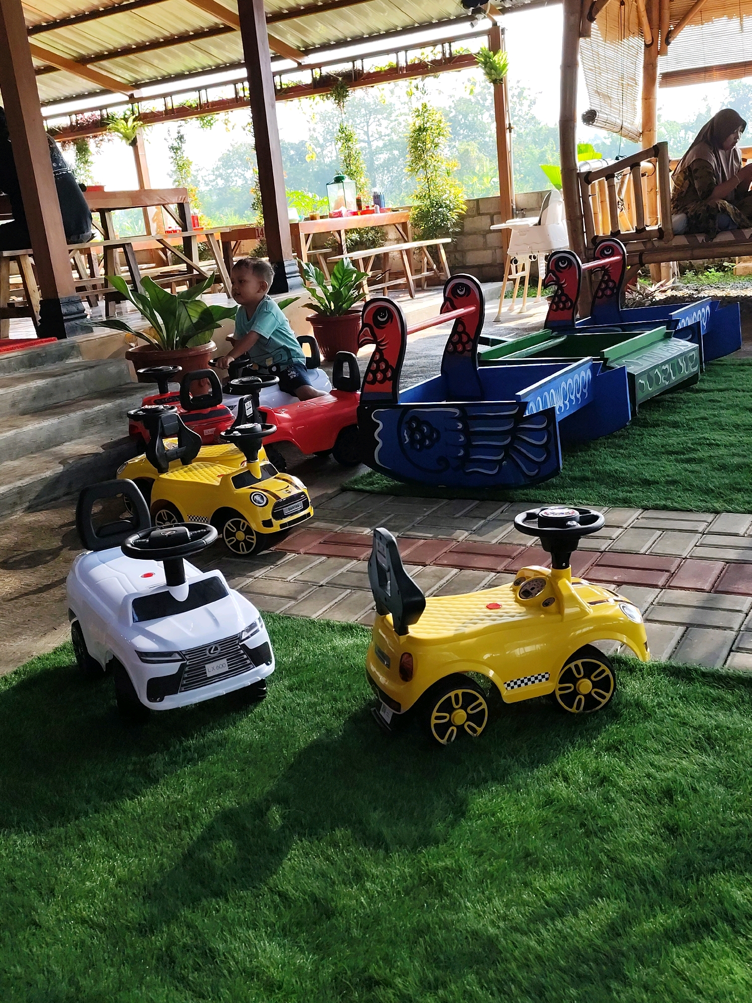 This image depicts an outdoor seating area at a restaurant in Klaten, Indonesia, likely a cafe or restaurant with a garden setting. In the foreground, several ride-on toy cars are arranged on a patch of artificial grass. These include a white Lexus-style car with "LX 600" on the license plate, a yellow Mini Cooper-style car, and a red car further back. A young child, wearing a blue shirt and dark shorts, is seated on the red toy car, looking to the left. Behind the toy cars, a row of decorative, colorful boat-like structures with bird figures are visible. The background shows a covered seating area with wooden tables and chairs, some with patrons seated. The overall ambiance suggests a family-friendly establishment, possibly a tourist spot or a popular local eatery in Klaten, a city known for its culinary offerings and cultural heritage. The natural light indicates it is daytime.