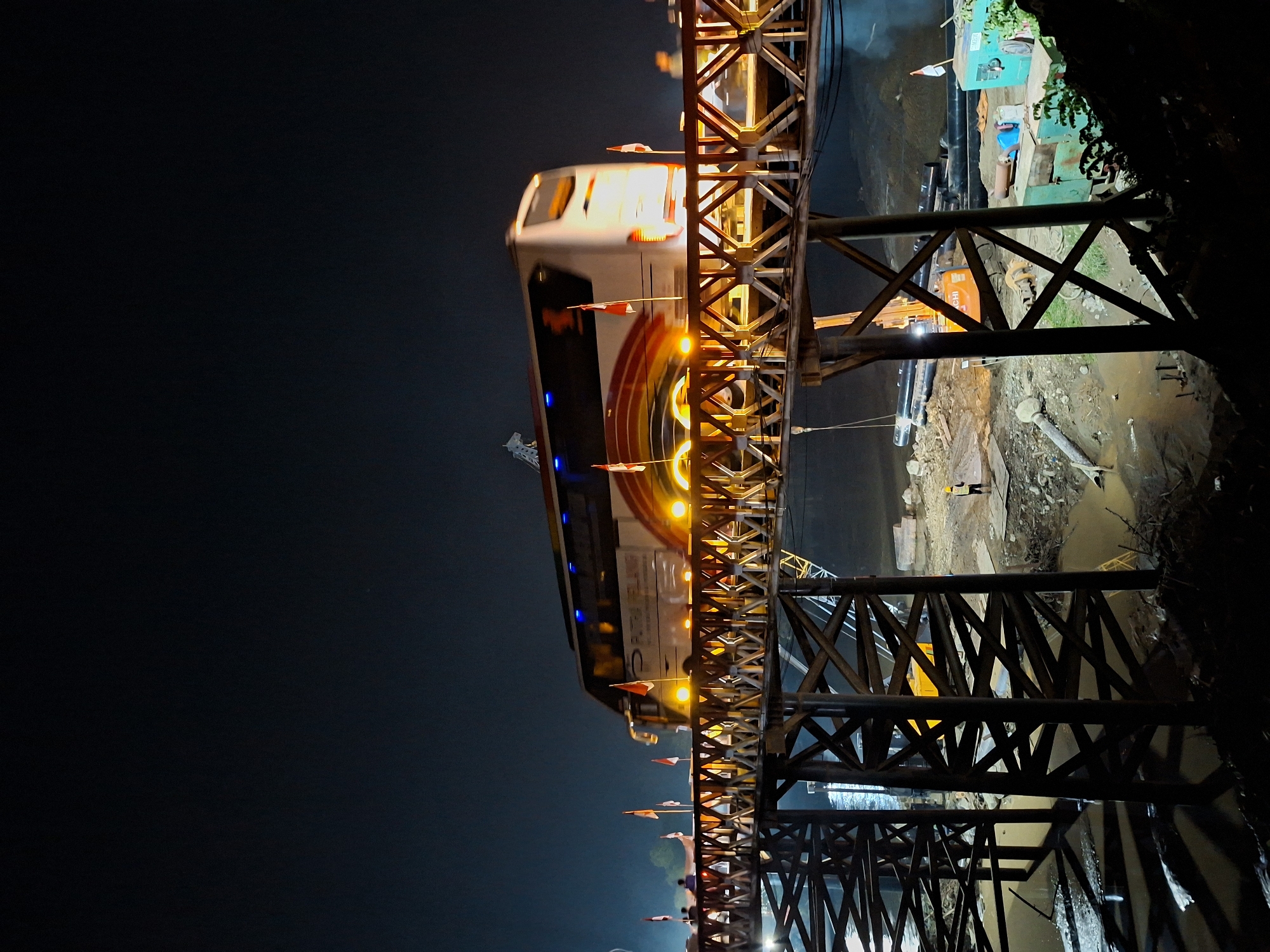 At night in Blang Mee, Indonesia, a train crosses a steel bridge, its lights illuminating the structure. Below the bridge, a construction site shows an excavator and workers, with the ground wet and muddy. The scene is a mix of infrastructure and industrial activity.