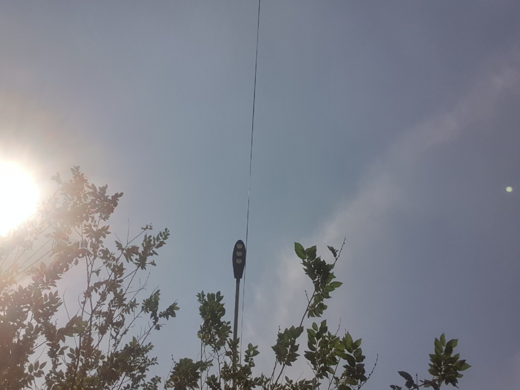 This is a high-angle, eye-level, medium shot of a streetlamp suspended on a wire against a hazy sky with sunlight filtering through from the top right corner. The streetlamp is dark gray and elongated, with three circular lights visible on its underside. It is positioned horizontally across the frame, with a thin wire extending from it to the right. The left side of the image is partially obscured by branches and green leaves from a tree, which are backlit by the sun, creating a silhouetted effect. The sky is a pale, overcast blue, with some faint clouds visible. The lower part of the sky has a slightly darker, more diffuse cloud formation. There are no people or animals visible in the image. The overall impression is one of an ordinary urban scene, likely taken on a moderately bright, hazy day. This scene is consistent with what might be observed in any urban environment, including the city of Shymkent, Kazakhstan.