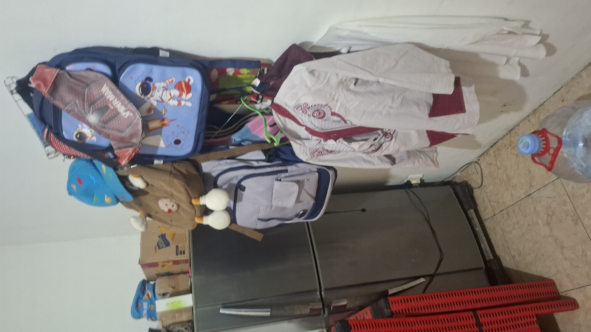The image depicts an indoor scene, likely a storage area or a hallway, located in the city of Cibinong Hilir, Indonesia. A grey refrigerator is positioned on the left side of the frame, partially obscured by a red plastic storage rack. Above and to the right of the refrigerator, several backpacks are hung on a rack attached to a white wall. These include a blue backpack with an astronaut design, a brown backpack with decorative elements, and a grey backpack. Clothing items, including a white shirt with red embellishments and a white garment, are also hanging on the wall. A large clear plastic water jug with a blue and red dispenser cap is visible on the floor in the lower right corner of the image. The floor is tiled with a light-colored pattern. The overall impression is one of a cluttered but functional domestic space.