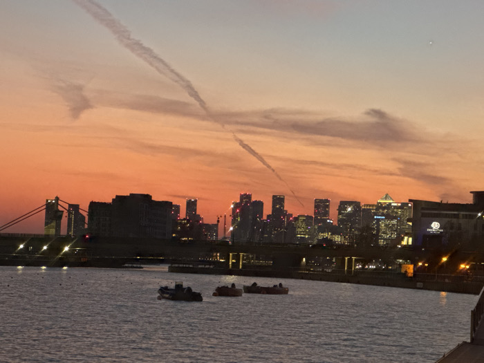 The image depicts a panoramic view of the Royal Victoria Dock in London, United Kingdom, at twilight, with the Canary Wharf skyline visible in the distance. The sky presents a gradient of deep orange and red at the horizon, transitioning upwards to lighter orange and pale blue. A distinct, winding white contrail from an aircraft diagonally traverses the upper-left portion of the sky, with a small, bright white point of light visible in the upper-right.
The foreground is dominated by the calm, dark water of the dock. Three small, dark, inflatable-style boats, possibly recreational pedal boats, are clustered in the mid-ground on the water. Behind these boats, an elevated, dark structure, appearing to be a bridge or pier, spans horizontally across the frame, illuminated by several distinct yellow-white lights.
The distant cityscape on the horizon is the illuminated silhouette of the Canary Wharf financial district. Numerous high-rise buildings, including the pyramid-topped One Canada Square, display illuminated windows against the darkening sky. To the far left, a large structure resembling a suspension bridge or industrial facility is partially visible. On the far right, a large building features a prominently illuminated "DoubleTree by Hilton" sign. No distinct individuals or specific human actions are discernible.