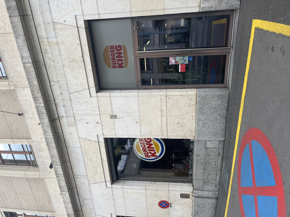 A daytime view captures the street-level facade of a Burger King restaurant in Genève, Switzerland. The building features a light-colored, possibly stone or marble, exterior with horizontal banding and a darker granite base.
The main entrance consists of glass doors framed in metal, situated to the right. To the left of the doors, a large window displays the modern Burger King "bun" logo, which is orange and white with "BURGER KING" text, partially visible through a frosted or tinted section of the glass. Below this, a smaller window showcases the older, circular Burger King logo with a blue circle, red "BURGER KING" text, and yellow accents, hanging inside the clear glass.
The asphalt street occupies the lower and right portions of the frame. A solid yellow line is painted on the asphalt, running parallel to the building. Prominently displayed on the asphalt is a large, circular red and blue symbol, indicating a "no parking" or "no stopping" zone, featuring a red border, a blue interior, and a red diagonal line. A smaller, identical "no parking/stopping" sign, a red circle with a blue background and a diagonal red line, is affixed to the building wall just below the lower Burger King window.
The lighting suggests it is daytime, likely overcast, as there are no harsh shadows. Reflections of other buildings and the sky are visible in the glass surfaces of the windows and doors. No people are present, and no specific activity is taking place. The only clearly readable text is "BURGER KING" on both logos.