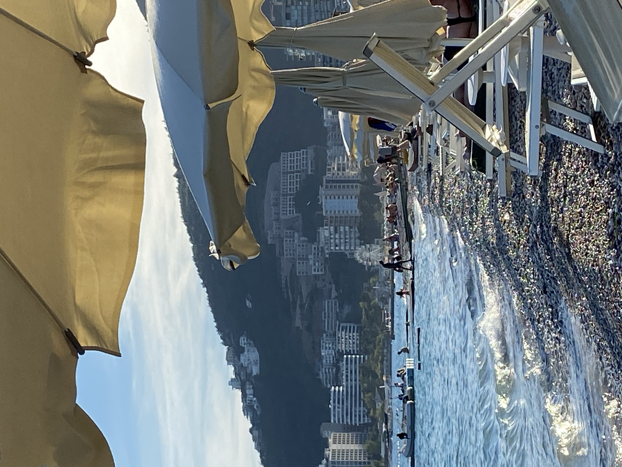 The image depicts a sunny day at a pebble beach in Budva, Montenegro. In the foreground, several white beach chairs and closed umbrellas are arranged on the shore, indicating a relaxation area. The sea, with gentle waves breaking on the shore, occupies the left side of the frame. In the middle ground, people are visible in the water and on small floating structures. Further back, a Ferris wheel is discernible on the coastline, and buildings are scattered across a verdant, hilly landscape that forms the backdrop. The sky is clear blue with some wispy clouds.