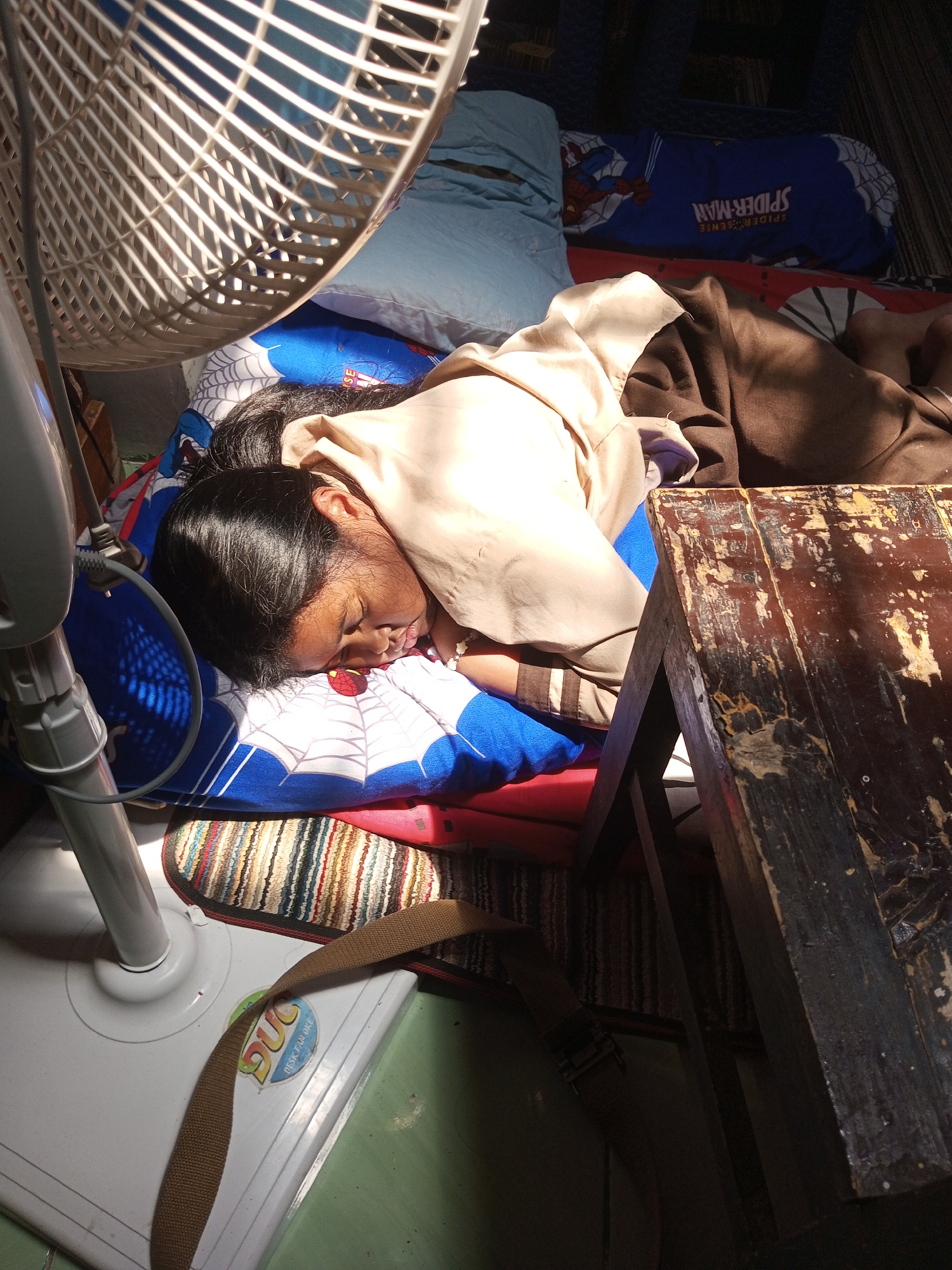 A young person is sleeping on a mattress on the floor. They are lying on their side, covered by a brown blanket and a light-colored shirt. The person is wearing a blue pillow with Spider-Man graphics. A white oscillating fan is positioned above and to the left of the person, casting shadows. A wooden table with a worn surface is in the foreground to the right. A woven mat and a strap are visible on the floor. The environment appears to be indoors, possibly a bedroom or living area, with lighting from a window or sunbeam. The scene suggests a moment of rest or sleep in a humble setting.