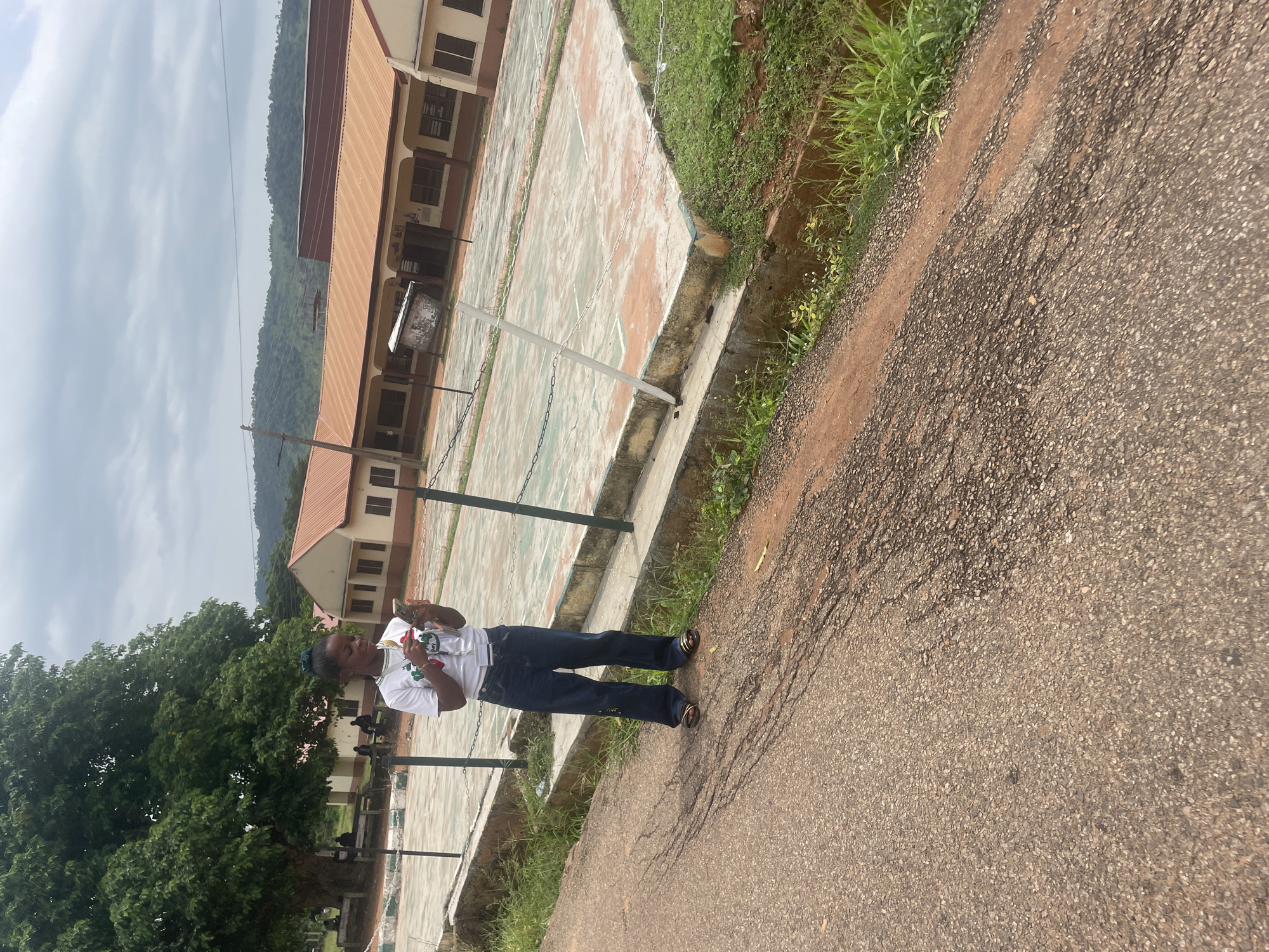 A young woman of African descent, wearing a white t-shirt, dark blue jeans, and sandals, stands on a paved road, looking at her phone. She has dark hair pulled back. Behind her is a chain-link fence, a concrete structure, a basketball court with faded markings, and a low-rise building with a brown roof. Green trees and a hillside are visible in the background under an overcast sky. The scene appears to be a school or recreational area in Lokoja, Nigeria.