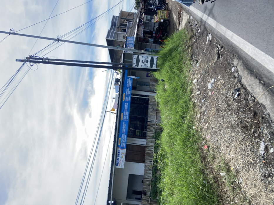 The image shows a street in Rancabali, Indonesia (latitude: -7.122, longitude: 107.428). There are several small shops and houses along the street. One of the shops has a sign that says "BIRA CELL" and advertises various services. Another shop has a sign that says "PANGKAS RAMBUT" and features a picture of a man with a haircut. There are power lines running across the top of the image, with two tall poles visible. The street is paved, and there are a few motorcycles parked near the buildings. The ground on the side of the road is covered in grass and gravel. A person is riding a motorcycle on the road, their legs can be seen, but their upper body is out of frame. The sky is partly cloudy.