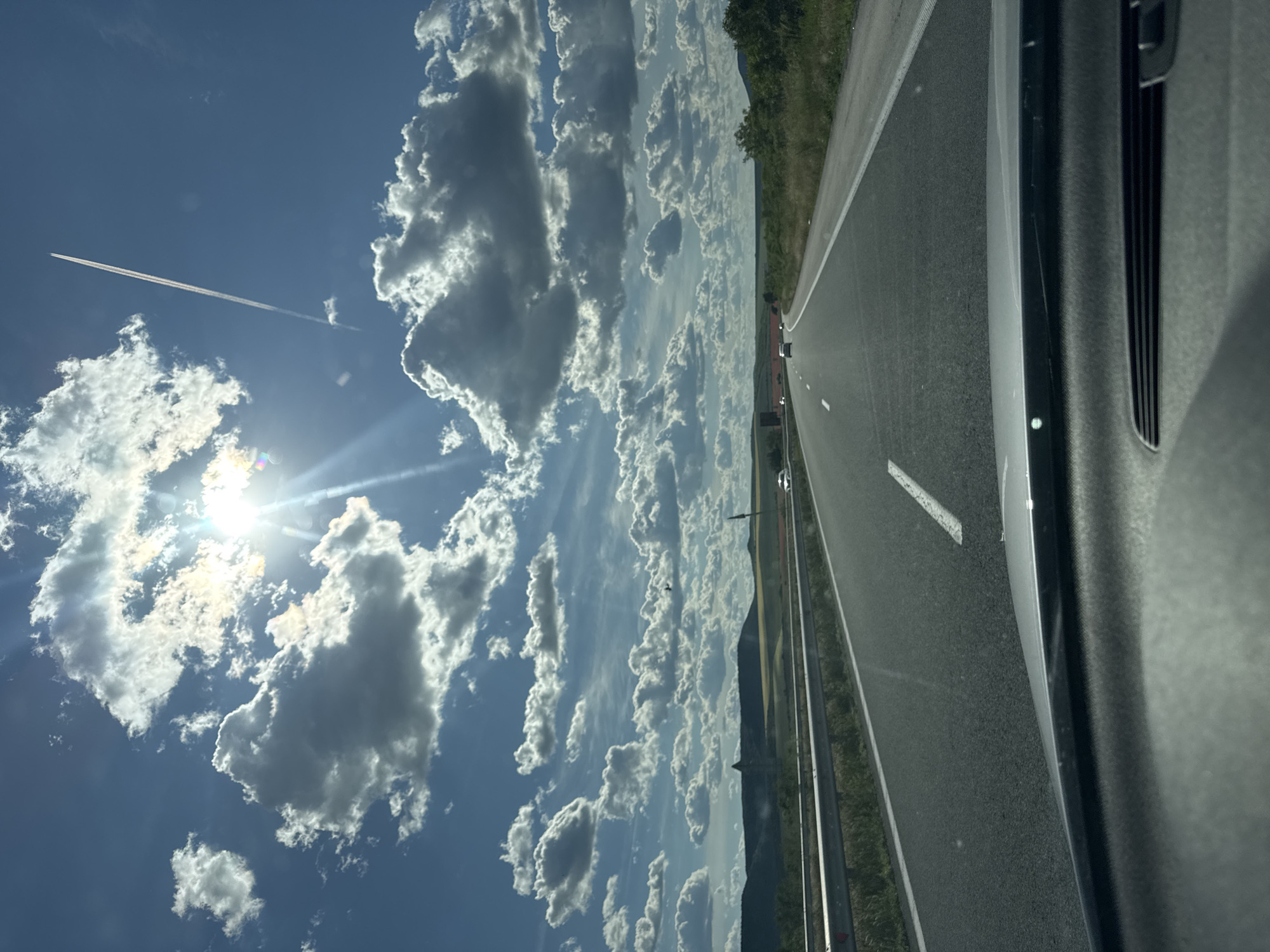 The image presents a view from inside a vehicle traveling on a highway near Zlatina, Bulgaria. To the left is a bright, sunlit sky filled with cumulus clouds and a contrail from an aircraft. The vehicle is on a paved road with lane markings and adjacent grassy areas leading to a distant treeline or hills. The interior of the car is visible on the right.