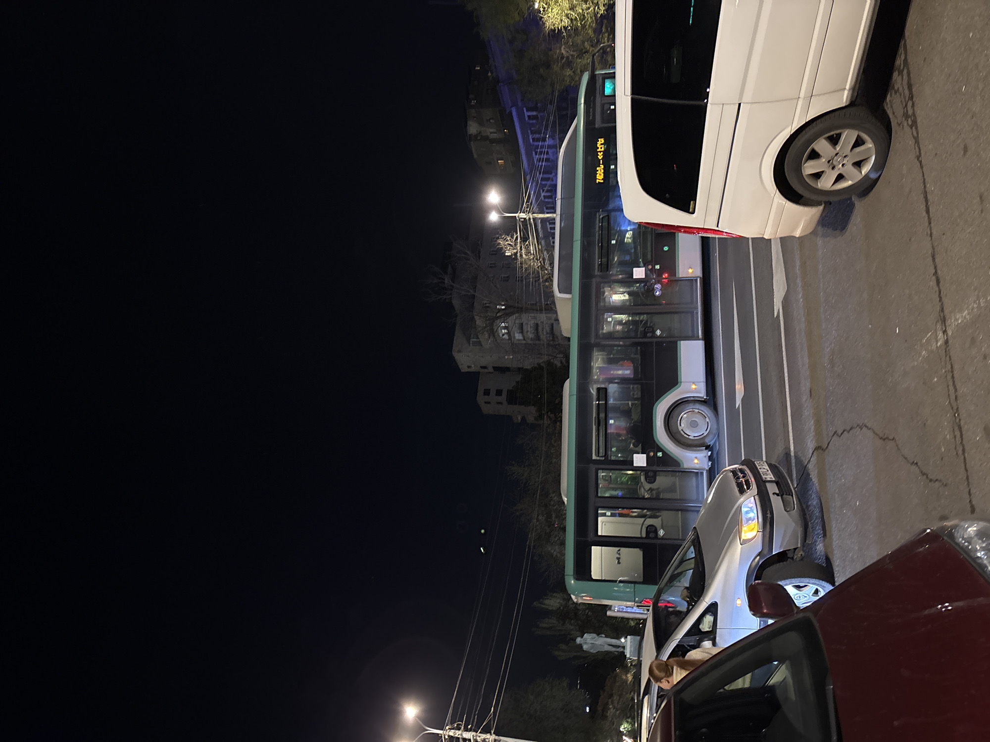 The image depicts a nighttime street scene in Yerevan, Armenia, showing a congested road with multiple vehicles, including a white van, a green and white bus displaying "Էրեբունի-Մեդիա", and other cars including a maroon car. The street is illuminated by streetlights. The scene appears to be in a busy location, based on the presence of multiple vehicles and the reflection of lights. Buildings can be seen in the background. A person with blond hair is seen in one of the cars.