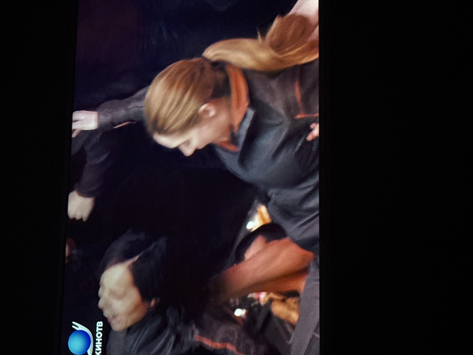 The image shows two individuals involved in a physical altercation. A woman with reddish-blonde hair in a ponytail, dressed in a dark top with orange trim, appears to be involved in a struggle with at least one other person. A man with dark hair is visible in the foreground, also wearing dark clothing. The setting is dimly lit, and the presence of a logo in the bottom left corner suggests the image may be a still from a video. There are no indications of a specific location in Mytishchi, Russia. The description focuses on observable details without speculation.