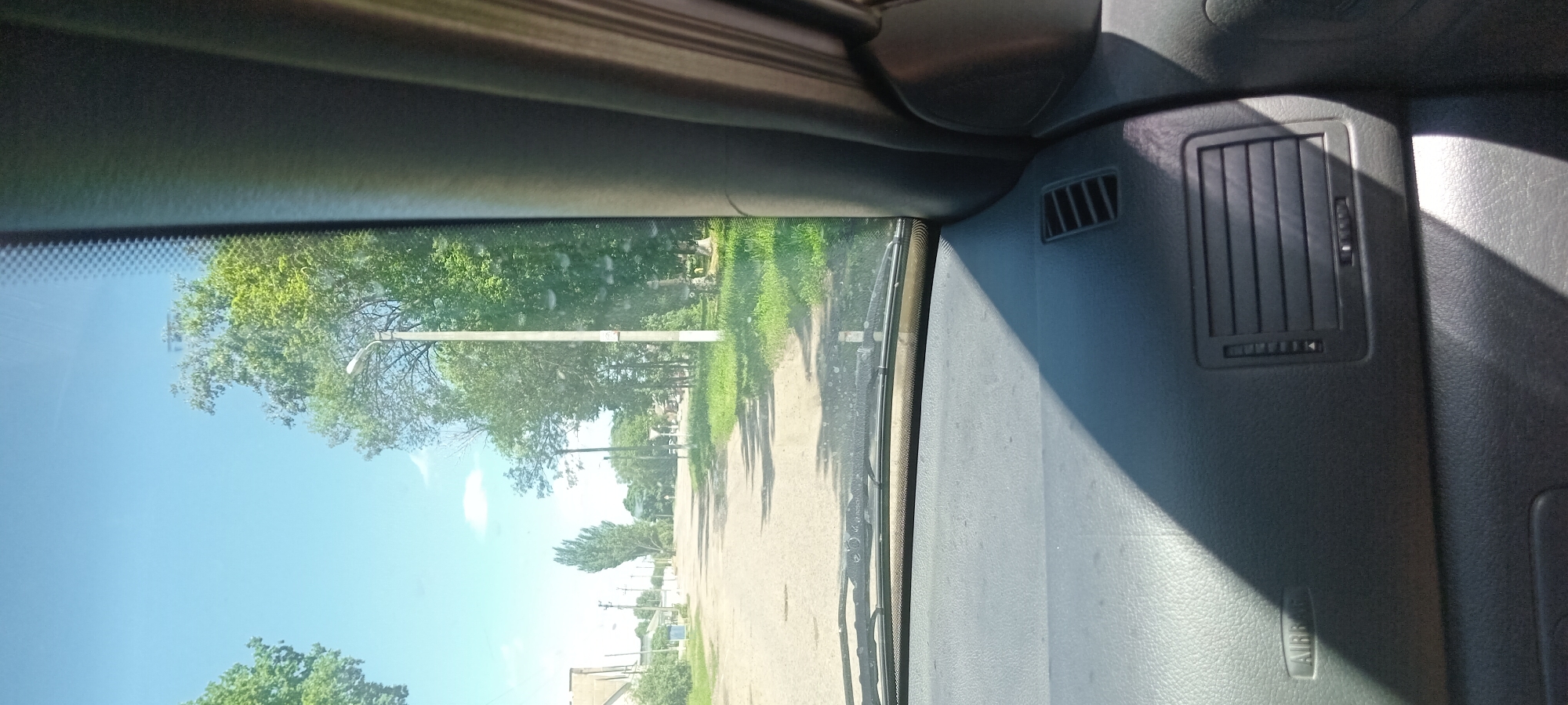 The image captures a view from the inside of a vehicle, looking through the windshield. The scene depicts a rural road lined with trees and grass under a clear blue sky. A lamppost stands on the right side of the road, and in the distance, there are buildings and more trees. The dashboard of the car is visible in the foreground, with an "AIRBAG" marking on the left side and air vents on the right. The image appears to be taken during daytime, with sunlight casting shadows on the dashboard. The location is identified as Koviahy, Ukraine.
