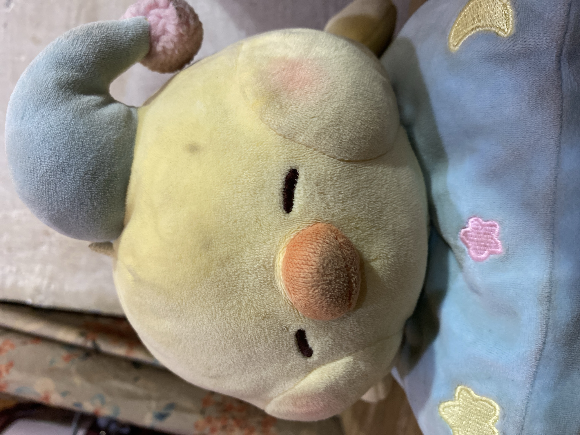 A close-up, waist-up, eye-level shot captures a yellow plush chick toy wearing a light blue, pointed sleeping cap with a pink pom-pom. The chick's eyes are closed, depicted as dark brown slits. Its beak is a textured orange, and its cheeks have a slight pink blush. The toy is resting on a blue fabric surface adorned with embroidered yellow and pink stars and a yellow crescent moon. The background consists of draped fabric with a floral pattern in muted colors and a plain, textured surface. The lighting is dim, casting soft shadows. This image was taken in Ta', Indonesia.