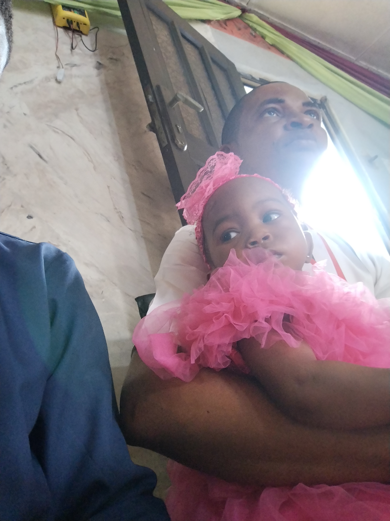 A man is holding a young child wearing a pink dress and headpiece. The man is looking upwards with his eyes slightly open. The child is looking to the left. A person wearing a blue sweater is to the left of the man. In the background, there is a marble wall with a yellow device attached, a dark-colored door with a silver handle, and green and maroon fabric draped across the ceiling. The location appears to be indoors. The city is unknown.