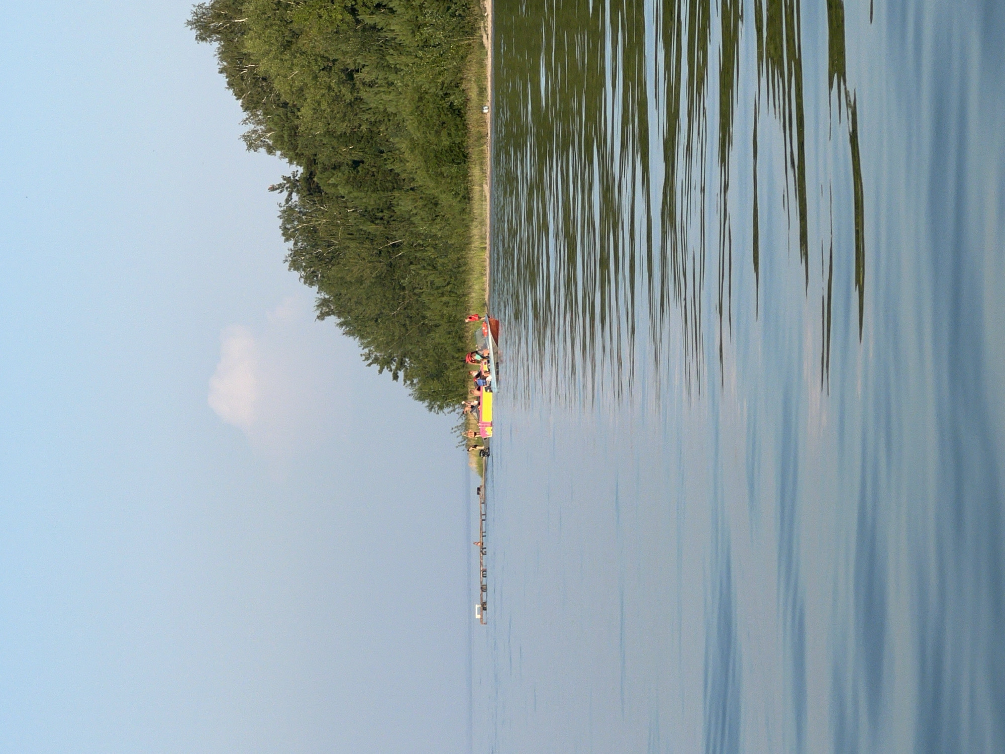 The image captures a serene scene on a body of water, likely a lake, with a lush green forest lining the distant shore. The water is calm, reflecting the pale blue sky and the verdant trees. In the middle ground, a wooden pier juts out into the water, with several figures visible standing on it. To the right of the pier, a brightly colored yellow and pink boat, possibly a pontoon boat, is anchored near the shore, carrying several people. The boat appears to be adorned with some form of advertisement or graphic. Further down the shore, more trees and vegetation are visible, with a few scattered individuals on the beach. The overall atmosphere is peaceful and recreational, suggesting a summer day by the water. The location is specified as Borovoe, Russia.