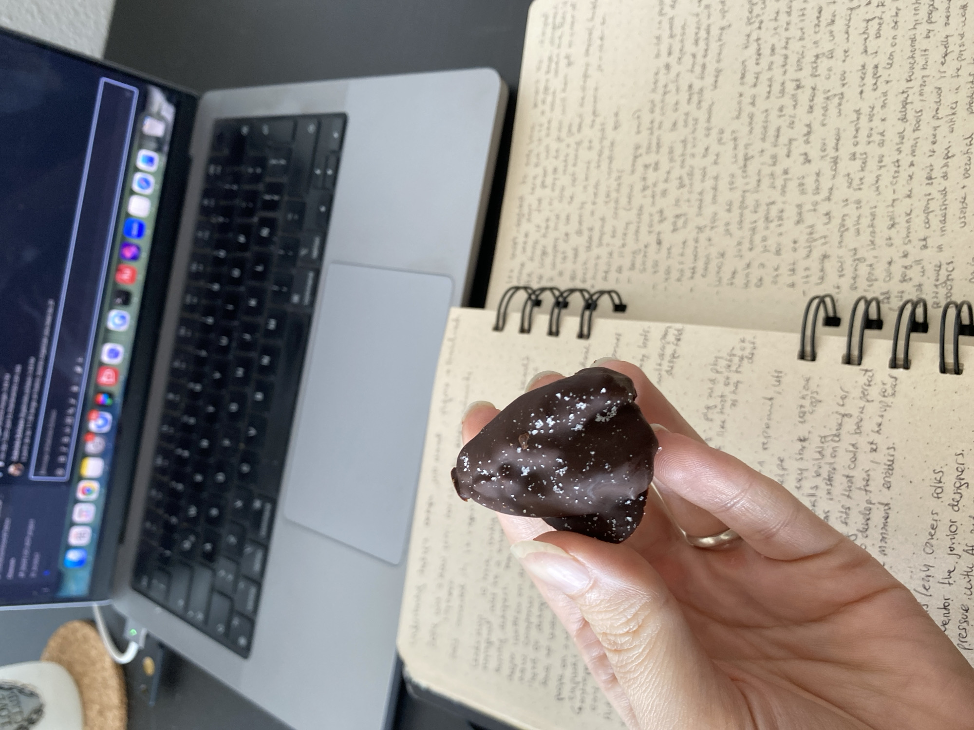 Here is a description of the image:
A hand holds a dark chocolate candy sprinkled with salt. The background shows a notebook filled with handwriting and a laptop. The laptop screen displays what appears to be a video call or presentation. The desk appears to be black or dark gray. The environment suggests a work or study setting.