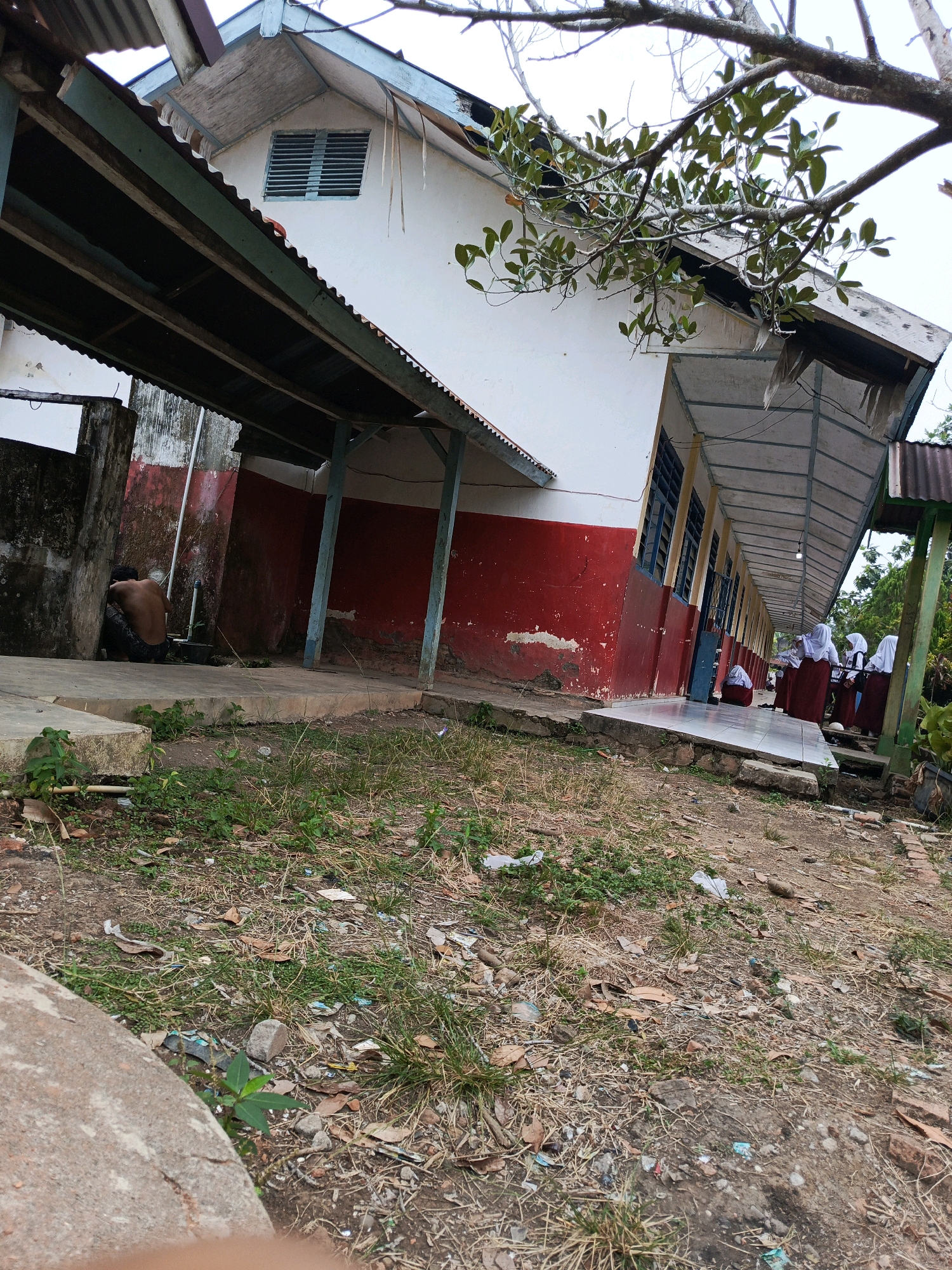 The image depicts the exterior of a simple, single-story building, likely a school, with an open-air covered walkway running along its length. The building has a white upper section and a red lower section, with blue trim visible on the roofline and supporting posts of the walkway. The walkway roof is corrugated metal. The ground in front of the building is unpaved, covered with dry grass, dirt, and scattered debris.
To the left, a bare-chested individual is crouched down next to a concrete structure. Further down the walkway, several figures, appearing to be young students in uniform (white tops and maroon skirts), are gathered, some standing and others seated. The building appears aged, with peeling paint on the red lower section.
The scene is outdoors, under an overcast sky. The environment suggests a somewhat neglected or rural setting within the city of Megang Sakti II, Indonesia. The overall impression is one of a functional, but not well-maintained, educational facility.