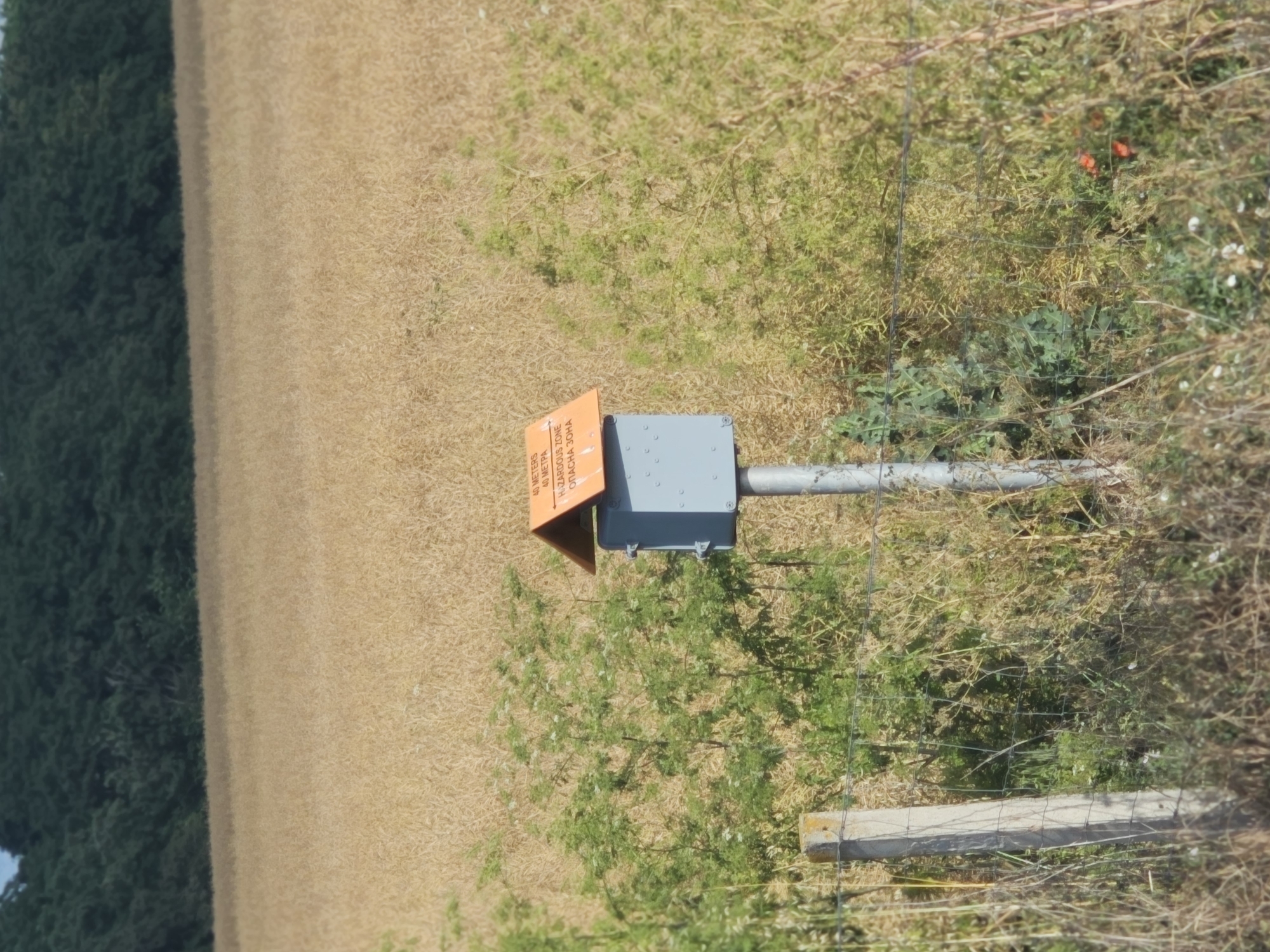 In Kolarovo, Bulgaria, a grey metal box with an orange, pitched roof sits atop a pole in a dry, grassy field. The box has a small, square flap and appears to be a weather station or monitoring device. Text on the orange roof reads "40 METERS 40 METRA HAZARDOUS ZONE ОПАСНА ЗОНА." A wire fence runs across the foreground, with a concrete post on the left. Beyond the field, a dense line of dark green trees forms the horizon. The sky is a pale blue.