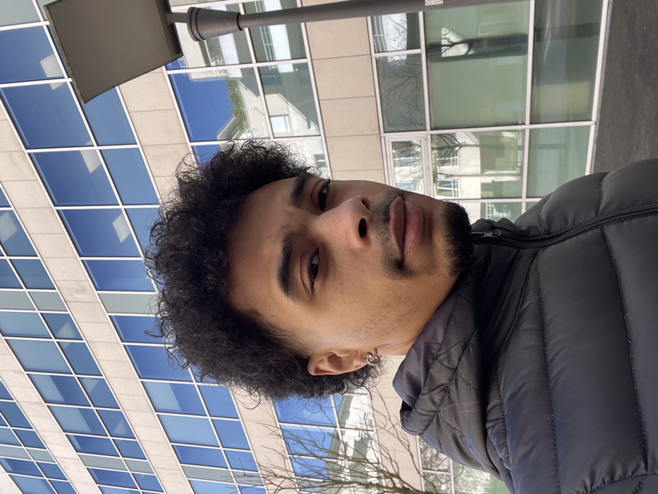 A young adult male is depicted in an outdoor urban environment within Montigny-le-Bretonneux, France. The subject has an olive skin tone, dark curly hair with faded sides, dark eyes, a mustache, and a goatee. He is wearing a black puffer jacket with a visible zipper. A silver-colored hoop earring is present in his left ear. He looks directly at the camera with a neutral facial expression. Behind the subject, a multi-story building with a light-colored, possibly beige, facade featuring numerous rectangular, reflective windows extends across the background. These windows reflect a clear blue sky, a lighter-colored building with balconies, and bare trees. To the right of the subject, a dark-colored streetlamp pole with a square-shaped light fixture is partially visible.
