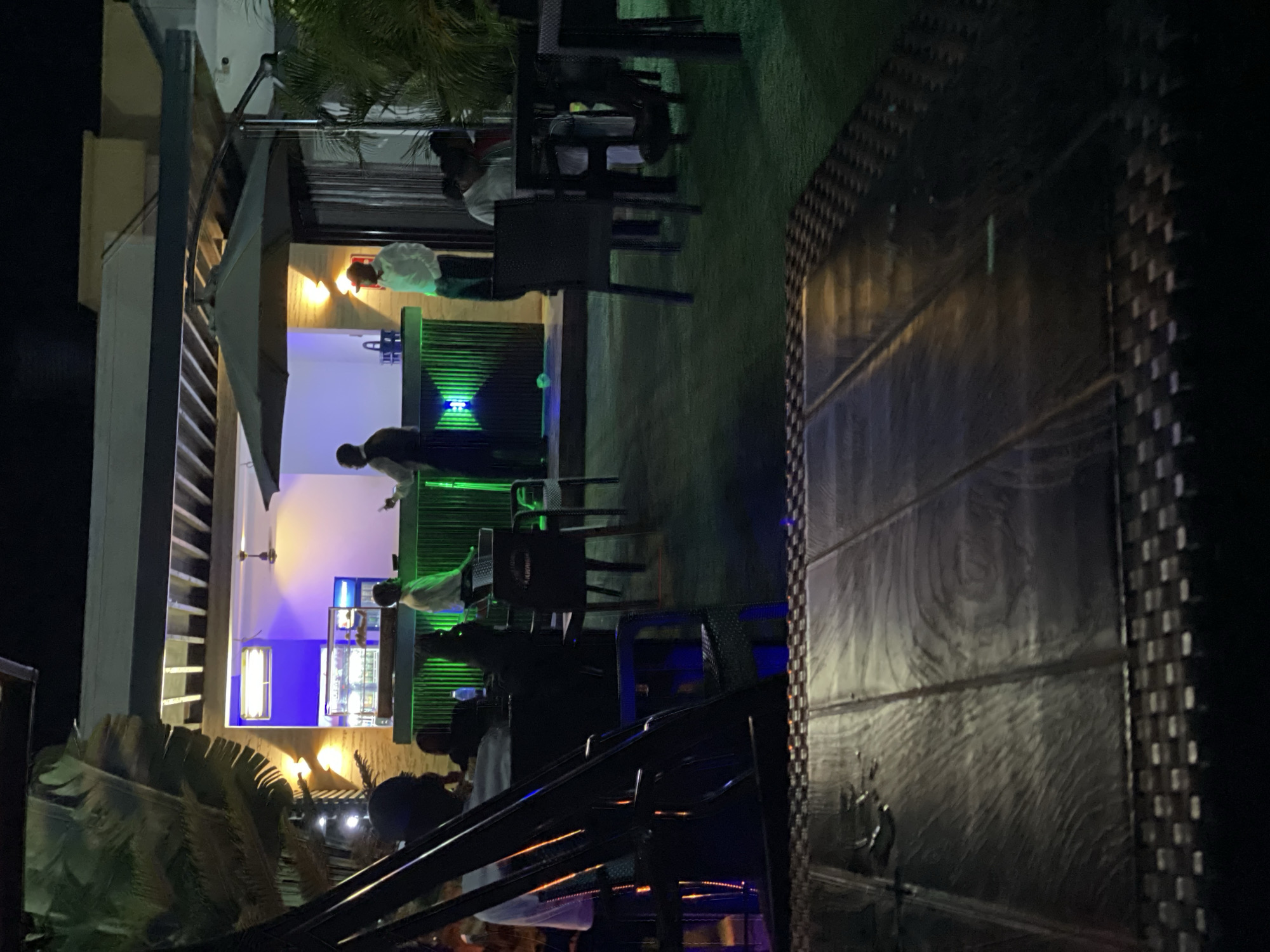 The image depicts an outdoor bar in Akwanga, Nigeria, at night. The bar features green and blue lighting. Several individuals are present, some appearing to be bartenders or staff behind the bar, while others are seated at tables. Tables and chairs are arranged in the outdoor area, with some covered by a canopy. A decorative wall or partition is visible in the foreground. Vegetation, including palm trees, is present in the vicinity of the bar.