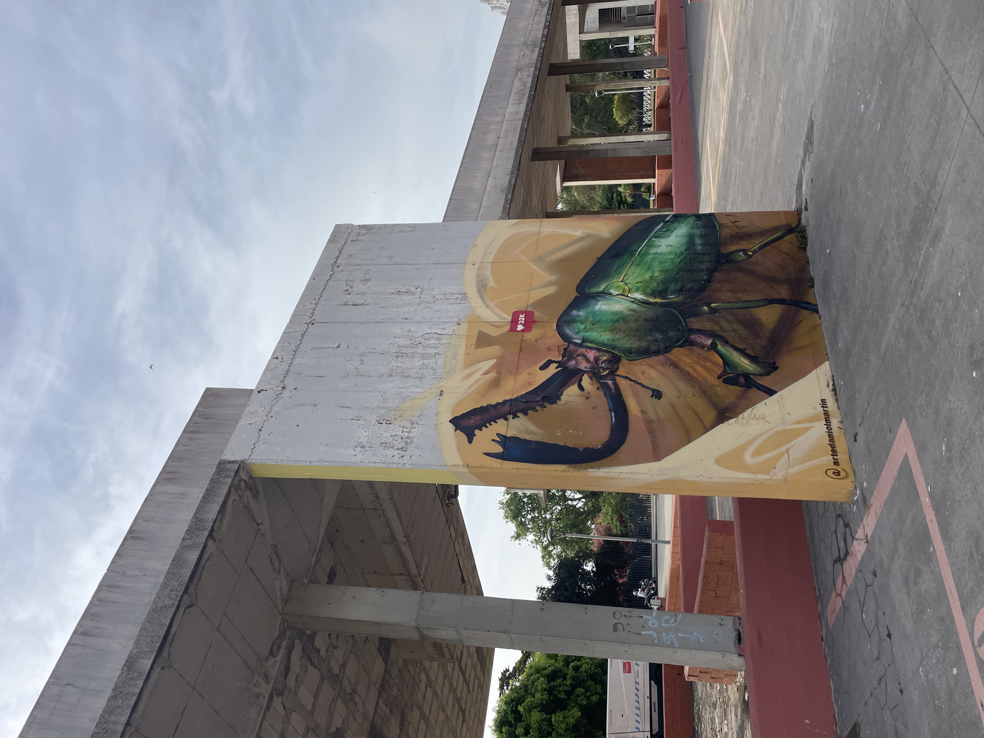 A large, vibrant mural of a green beetle is painted on a concrete wall in La Línea de la Concepción, Spain. The beetle is depicted in detail, with its iridescent green exoskeleton and large, segmented legs. The mural is set against a warm, golden background that resembles an abstract representation of a flower or leaf. A small heart icon with "32k" inside is superimposed on the mural, suggesting a digital endorsement. Below the mural, the artist's handle "@artedanielmartin" is visible. The concrete wall is weathered, with some signs of wear and tear.
The mural is located outdoors, in what appears to be a public space. In the background, there is a covered walkway supported by concrete columns, reminiscent of a bus station or a public building's architecture. Trees and greenery are visible beyond the walkway, suggesting a park or an urban area with some vegetation. The sky above is partly cloudy. To the left of the mural, a white bus or service vehicle is parked, and a person on a motorcycle is seen in the distance. The ground is paved with concrete and marked with pink lines, possibly indicating a sports court or designated area. The overall impression is a blend of urban infrastructure and public art, set under a daytime sky.