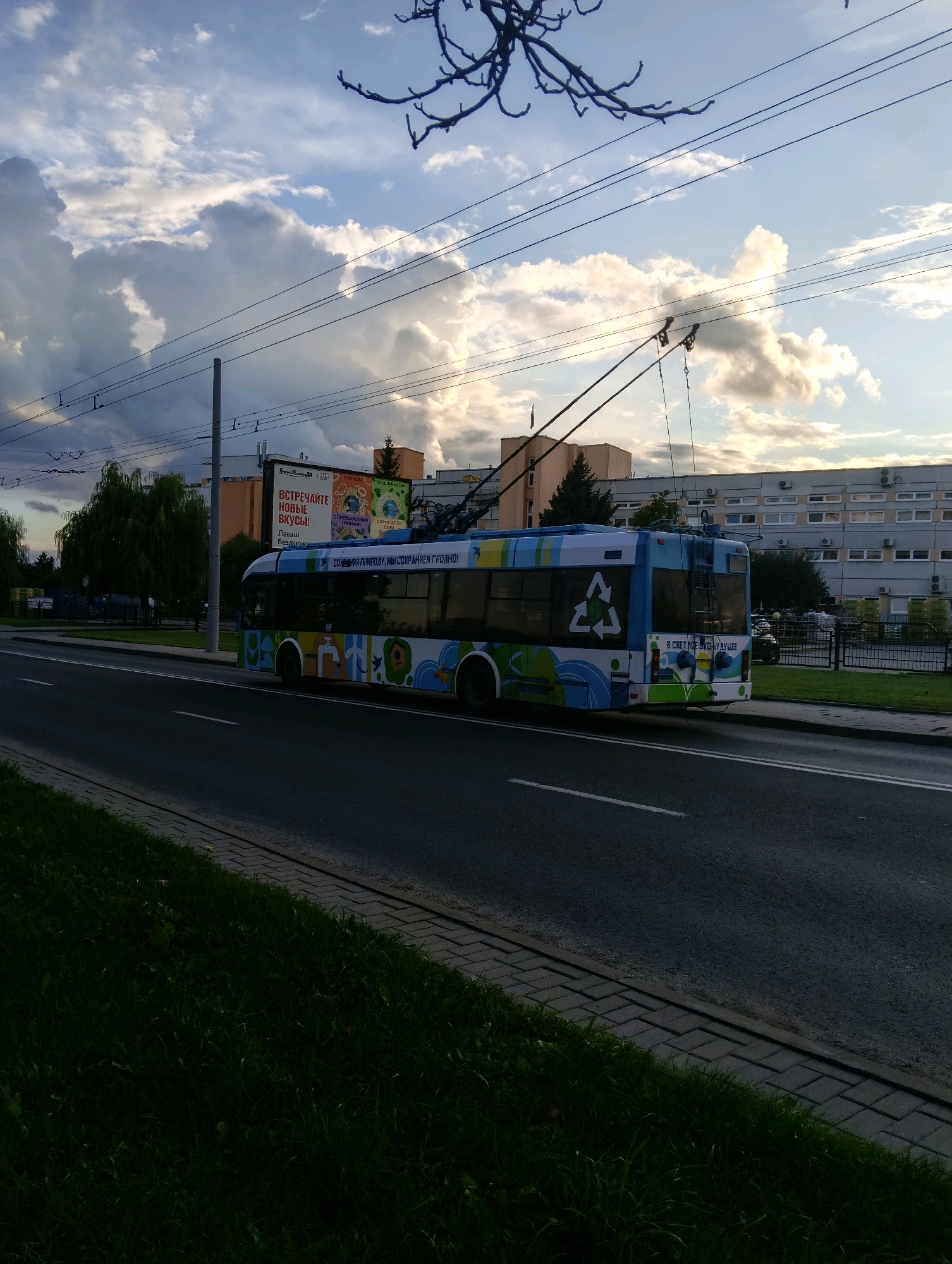 A trolleybus with a colorful design, featuring a recycling logo and a Belarusian slogan, drives down a street in Hrodna, Belarus. The trolleybus is blue and white with green and yellow accents, and the slogan "В СВЕТЛОЕ БУДУЩЕЕ" (Towards a bright future) is visible on its side. It is connected to overhead power lines, indicating it is in operation.
The trolleybus is driving on a paved road with a white lane marking. To the left of the road, there is a grassy area with a paved walkway. In the background, there are buildings, trees, and a large billboard with advertisements. The sky is partly cloudy, with dramatic clouds casting shadows on the scene.
The billboard features text in Belarusian, including "ВСТРЕЧАЙТЕ НОВЫЕ ВКУСЫ!" (Meet new tastes!) and "С ПРОБЛЕМЫ ГРУДНО!" (With problems of Hrodna!). The overall impression is a typical urban scene in Hrodna, showcasing public transportation and local advertising. The image was taken during daylight hours, likely in the late afternoon or early evening, judging by the lighting and the colors of the sky.