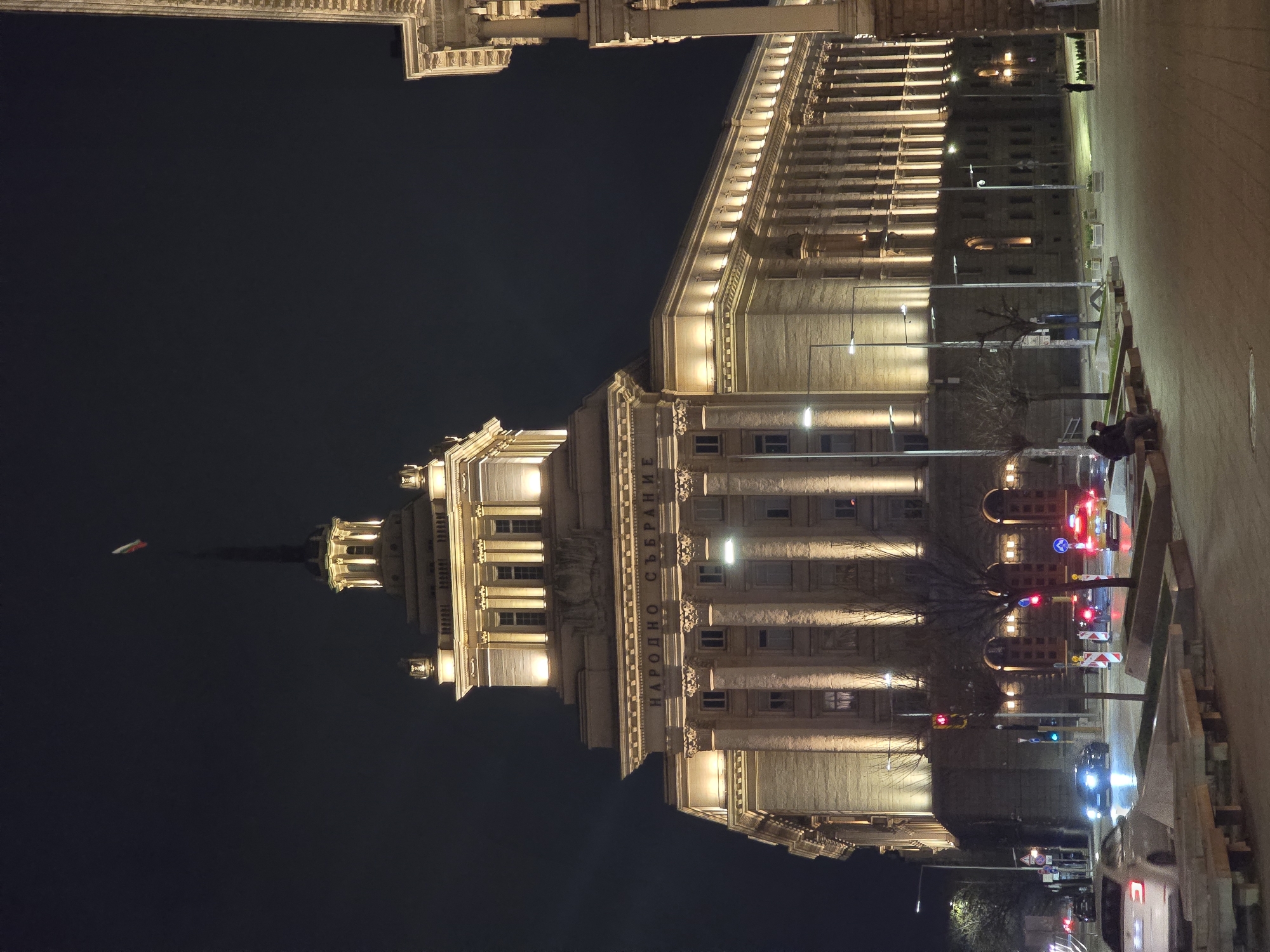 An outdoor night shot shows the National Assembly of Bulgaria building in Sofia, Bulgaria. The illuminated building is primarily light beige and white with a flag atop the highest tower. Columns frame the building, which has visible signage in Cyrillic text. To the right, a sidewalk area and street scene is visible, showing traffic lights, street lights, cars, construction barriers, and two people sitting on a bench. The sky above the building is a dark black.