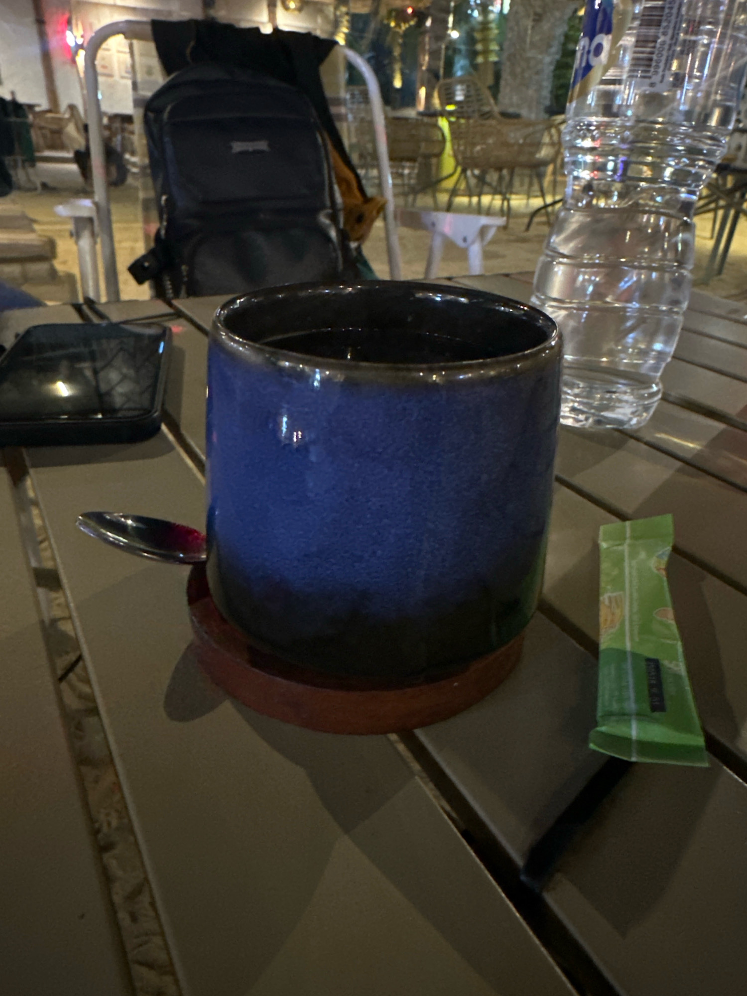 In Mojokerto, Indonesia, an outdoor table at a cafe setting displays a dark blue ceramic mug with a wooden coaster. A silver spoon rests on the edge of the coaster, partially submerged in the mug's dark liquid. To the right of the mug, a green packet of what appears to be sweetener or coffee creamer is placed on the table. A partially visible plastic water bottle is positioned behind the mug. In the background, a black backpack is slung over a white metal chair, with other cafe furniture and patrons visible in the dimly lit outdoor environment.