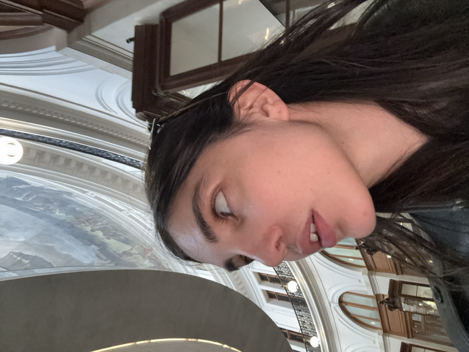 A woman with dark hair and fair skin is tilted to the side. She gazes downward with partially open lips. The backdrop reveals intricate architectural details: arched windows, ornate railings, and a ceiling mural depicting various figures and landscapes. A prominent circular structure with lighting is also visible. The setting appears to be an indoor architectural space of historical or cultural significance.