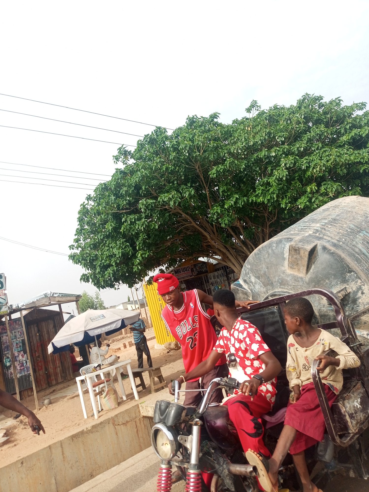 The image depicts a street scene in what appears to be a city in Nigeria, possibly Tempure, Nigeria. Three individuals are the primary focus: a young man wearing a red Chicago Bulls jersey and a red cap is standing next to a motorcycle rickshaw. Seated on the motorcycle is another young man in a red and white patterned shirt and red pants. A young girl is seated in the rickshaw section. The young man on the motorcycle is holding the handlebars. In the background, there are people seated at a table under an umbrella in front of a small shop. A large tree dominates the upper portion of the frame. Electrical wires are visible against the sky. A large water tank is attached to the back of the rickshaw. The general environment suggests an urban or semi-urban setting with dusty roads and simple structures.