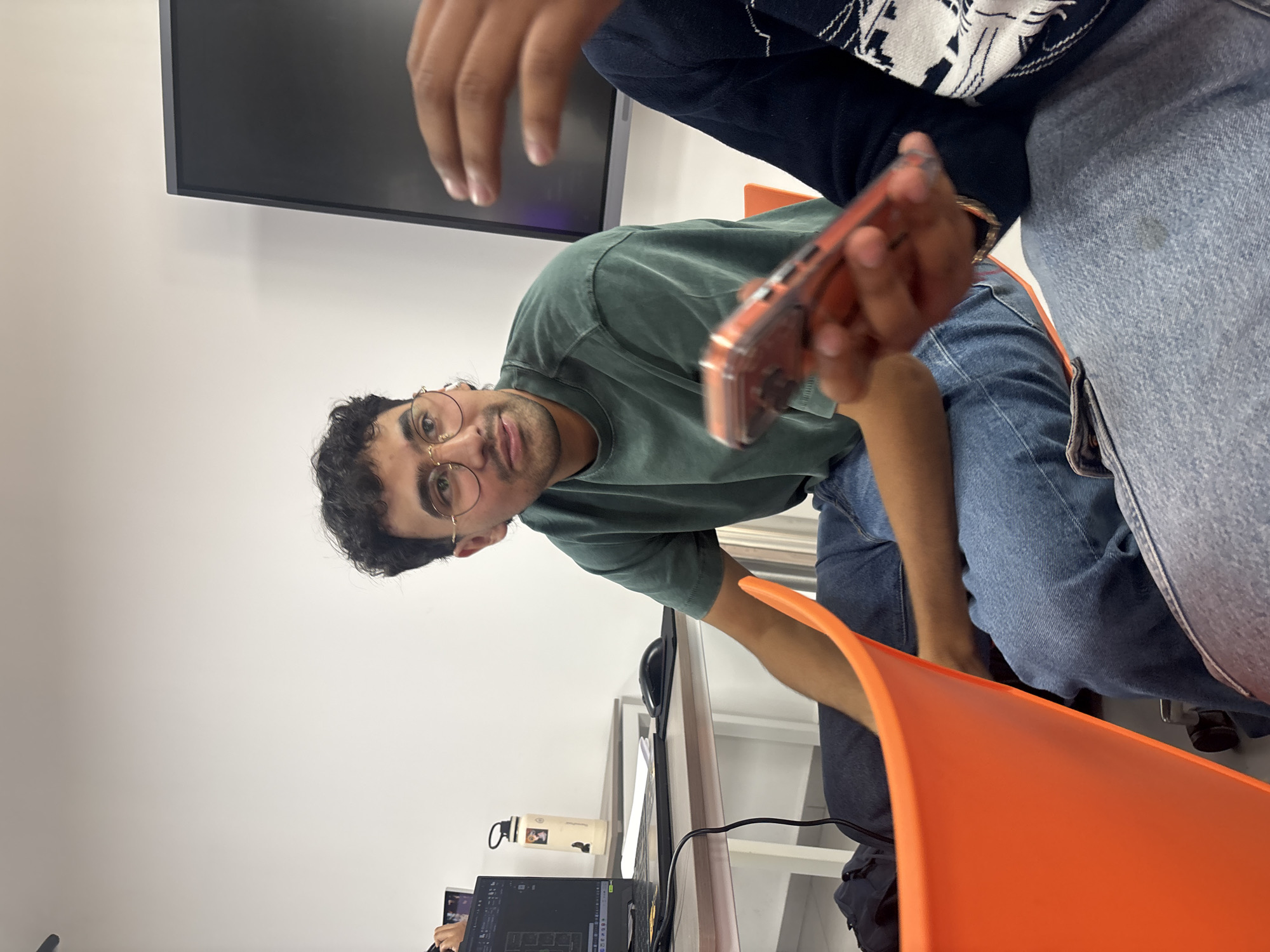 The image shows three people indoors. One person, wearing a green t-shirt and glasses, is looking towards the camera, holding a mobile phone. The other two individuals are seated; one wears denim and a dark sweater, while the other is wearing denim, with only their arm and lower leg showing. They are seated on an orange chair. In the background, there's a desk with a computer monitor, keyboard, and mouse on it. The setting appears to be a workspace. The location is San Andrés Cholula, Mexico.