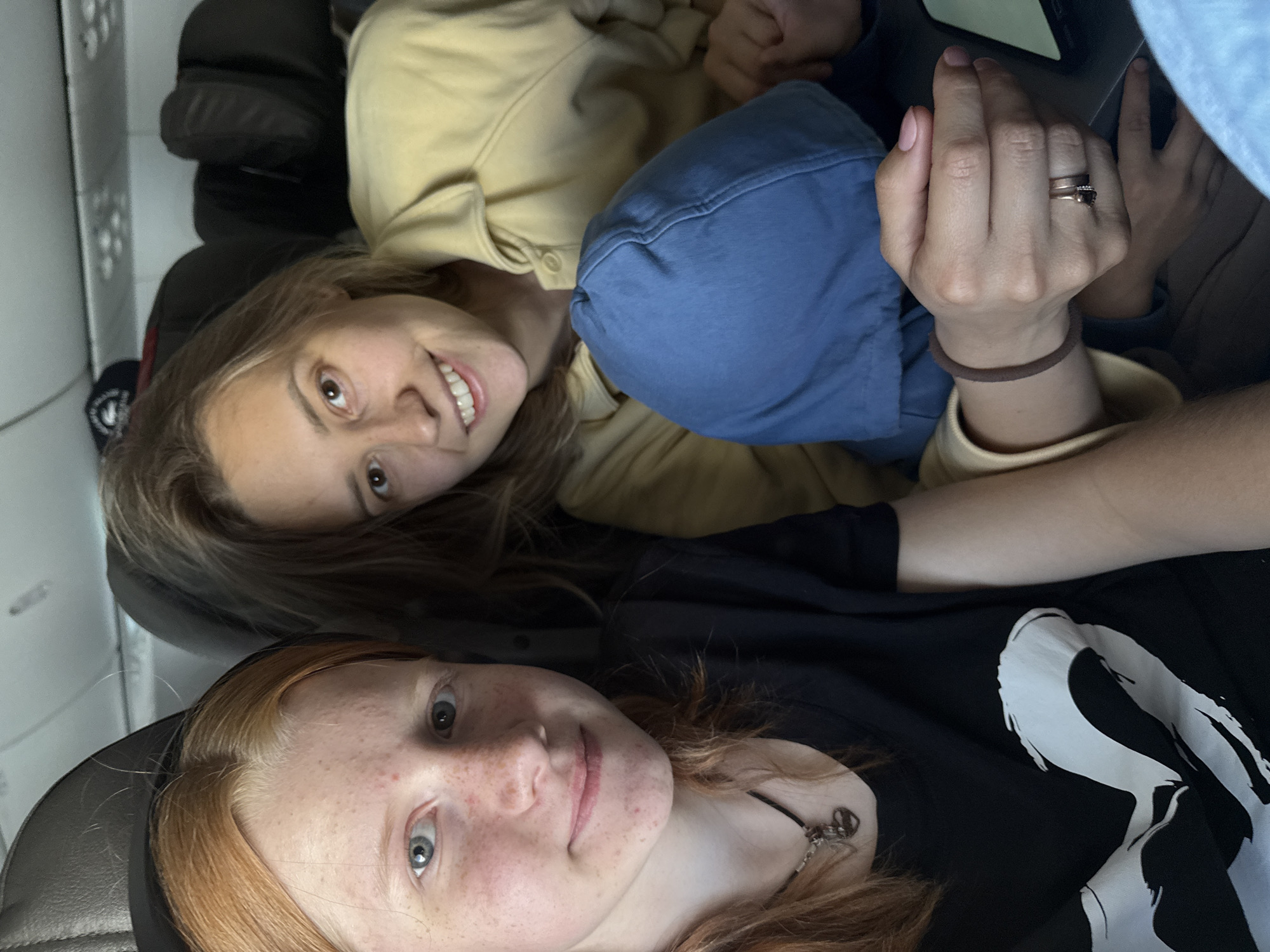 The image is a rotated, close-up shot featuring two young women, likely a selfie, taken in a dimly lit indoor environment, possibly within a vehicle.
In the foreground, a woman with reddish-brown hair and freckles on her face looks directly at the camera with light-colored eyes and a subtle smile. She is wearing a black t-shirt with a large, abstract white graphic design on the chest, and a necklace with a peace sign pendant.
Behind her, a second woman with light brown hair smiles broadly, revealing her teeth, and gazes towards the camera. She is dressed in a light yellow or beige long-sleeved top, with a blue garment, possibly a hoodie, pulled up around her head, partially covering her face. Her visible right hand displays two rings on her ring finger (one a simple band, the other with a dark stone) and a brown hair tie or bracelet on her wrist.
Both women appear relaxed and cheerful. The background consists of dark, upholstered surfaces, suggesting vehicle seating. The overall low lighting indicates it might be evening or night. No explicit text is visible, apart from the abstract graphic on the foreground woman's shirt.