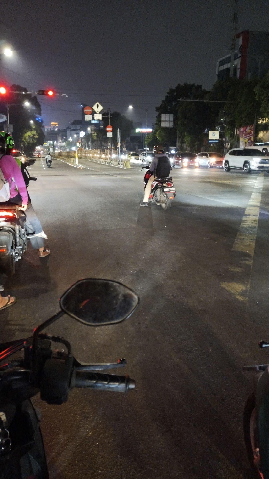 The image captures a nighttime street scene in South Jakarta, Indonesia, likely at an intersection. Several motorcycles and cars are present on the wet asphalt road, suggesting it might have recently rained or is about to. Traffic lights are visible, with one appearing to be red. Road signs, including a cautionary sign with an exclamation mark inside a diamond and a circular sign with a red border, are also present.
In the foreground, the handlebars and a portion of a motorcycle's mirror are visible, indicating the photo was taken from a motorcycle. In the mid-ground, a person in a pink jacket is seated on a motorcycle, their legs visible. Further ahead, another person on a motorcycle, wearing a grey jacket and a backpack, is also visible. Several cars are on the road, some with their headlights on. In the background, buildings and trees line the street, illuminated by streetlights and vehicle headlights. The overall atmosphere is that of a typical urban traffic scene at night.