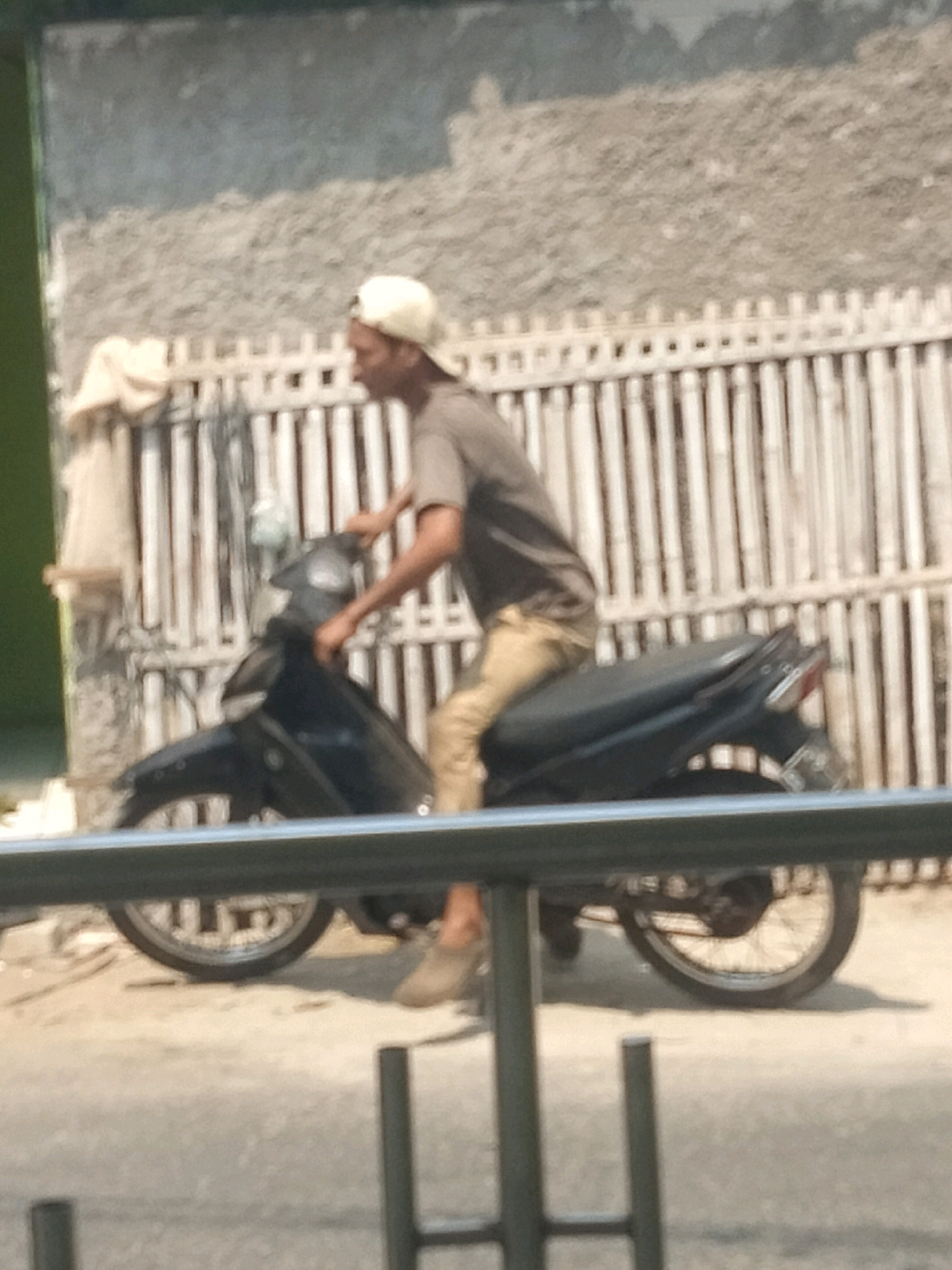 A person wearing a beige cap and a light brown t-shirt is riding a dark-colored motorcycle in Pisangan Jaya, Indonesia. The person is positioned in the center of the frame, with their body turned slightly to the left. Their right hand is on the handlebars, and their left hand appears to be near the controls. The motorcycle is a scooter-type, with a black body and visible wheels. Behind the motorcycle, there is a white wooden fence, and beyond that, a textured gray wall. The ground is a paved surface, likely a road or sidewalk, with some debris visible. The overall scene is outdoors, under what appears to be bright daylight, casting shadows. A metal railing runs across the lower portion of the image, partially obscuring the motorcycle's wheels.