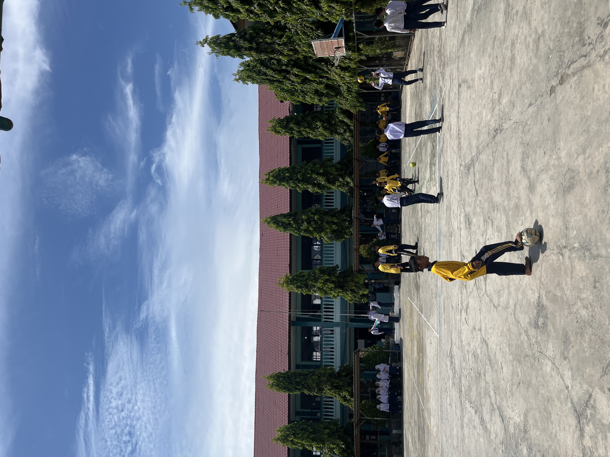 The image depicts a lively scene at a school in Lhokseumawe, Indonesia, likely during a school day or event. The central focus is an outdoor court area with a concrete surface marked with white lines, suggesting it's used for sports. In the foreground, a young person in a yellow long-sleeved shirt and dark pants is playing with a soccer ball, poised to kick it. Several other students, mostly boys, are engaged in various activities across the court. Some are playing a game that involves a yellow ball, possibly volleyball, near a basketball hoop and backboard. Others are standing or walking, observing the activities.
In the background, a two-story school building with a red-tiled roof and teal-colored walls is visible. The building features a prominent balcony with railings. Tall, slender trees are planted in a row along the front of the building, framing the scene. A group of students in white uniforms are seated on a bench under a shaded structure to the left of the building. The sky above is a vibrant blue, dotted with wispy white clouds, indicating fair weather. The overall atmosphere is one of youthful energy and camaraderie.