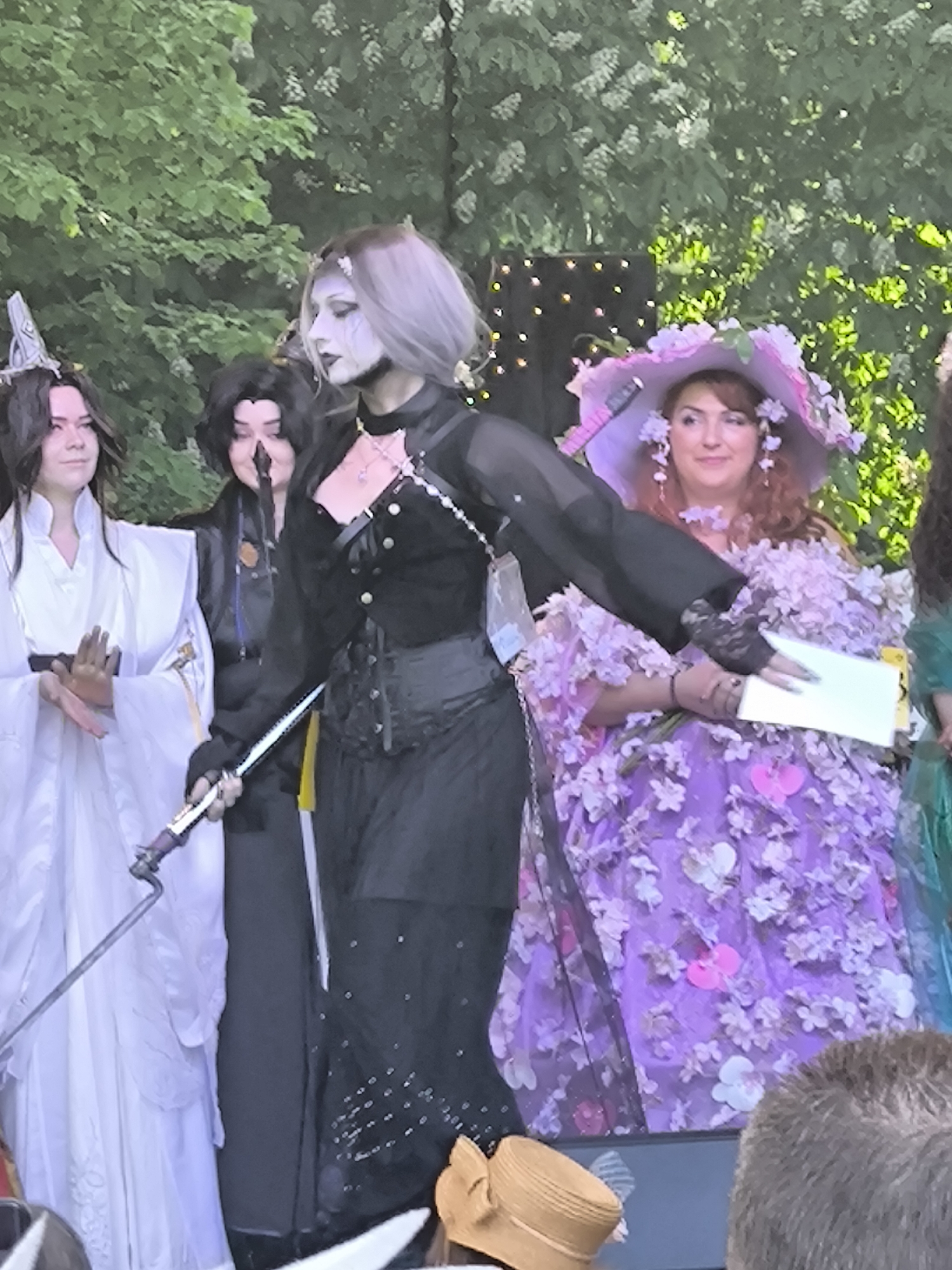 The image depicts a group of cosplayers on an elevated stage, possibly during a competition or event. The setting is outdoors, suggested by the green foliage and trees in the background.
From left to right, the individuals are:
1. A cosplayer in a white gown with a distinctive silver headpiece.
2. A cosplayer in black clothing, holding a prop microphone.
3. A cosplayer with light gray hair and white face makeup, wearing a black outfit, including a corset and cape, holding a sword.
4. A cosplayer wearing a purple dress covered with artificial flowers and a matching hat.
5. Part of another individual in a green dress is also visible on the far right.
The cosplayers are standing on a stage that has at least three steps leading to it. A golden colored hat is located near the base of the stage. The person in the purple dress seems to be holding a piece of paper.
The photo's quality suggests it was taken with a mobile phone or a camera with limited zoom capabilities. The location is not evident within the photo; the city of Riga, Latvia, cannot be determined from the visual information provided.