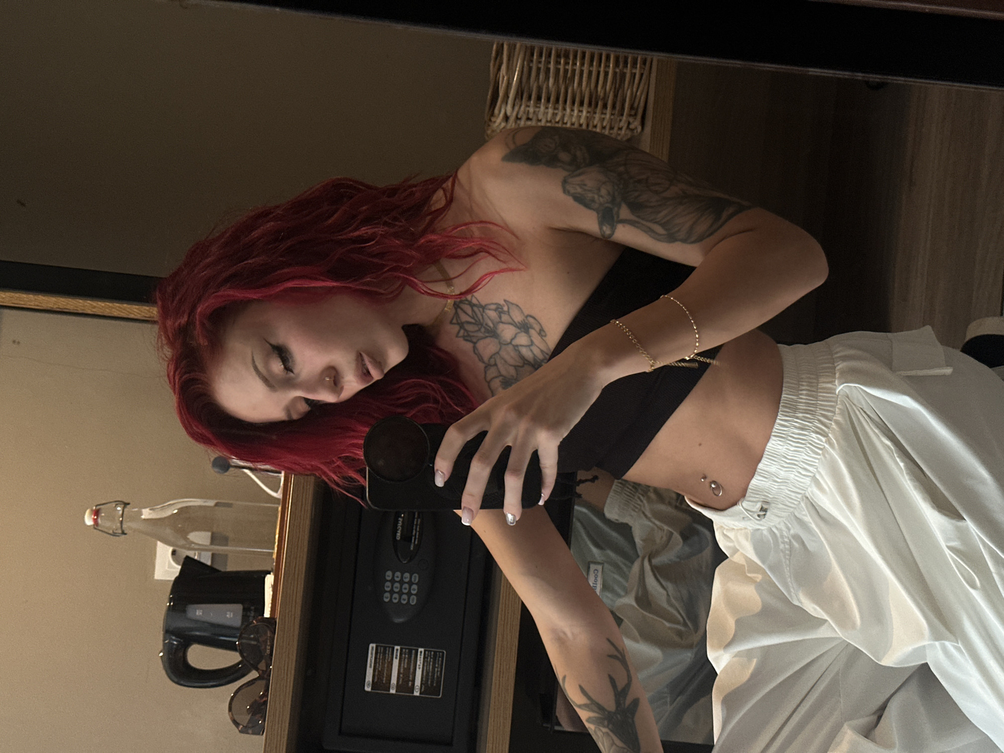 A woman with red, curly hair is taking a selfie in front of a mirror in a room. She is wearing a black tube top and white pants. She has tattoos on her arms and chest. A safe, a bottle, a kettle, and sunglasses are on a shelf behind her. The location is Unknown, Indonesia.
