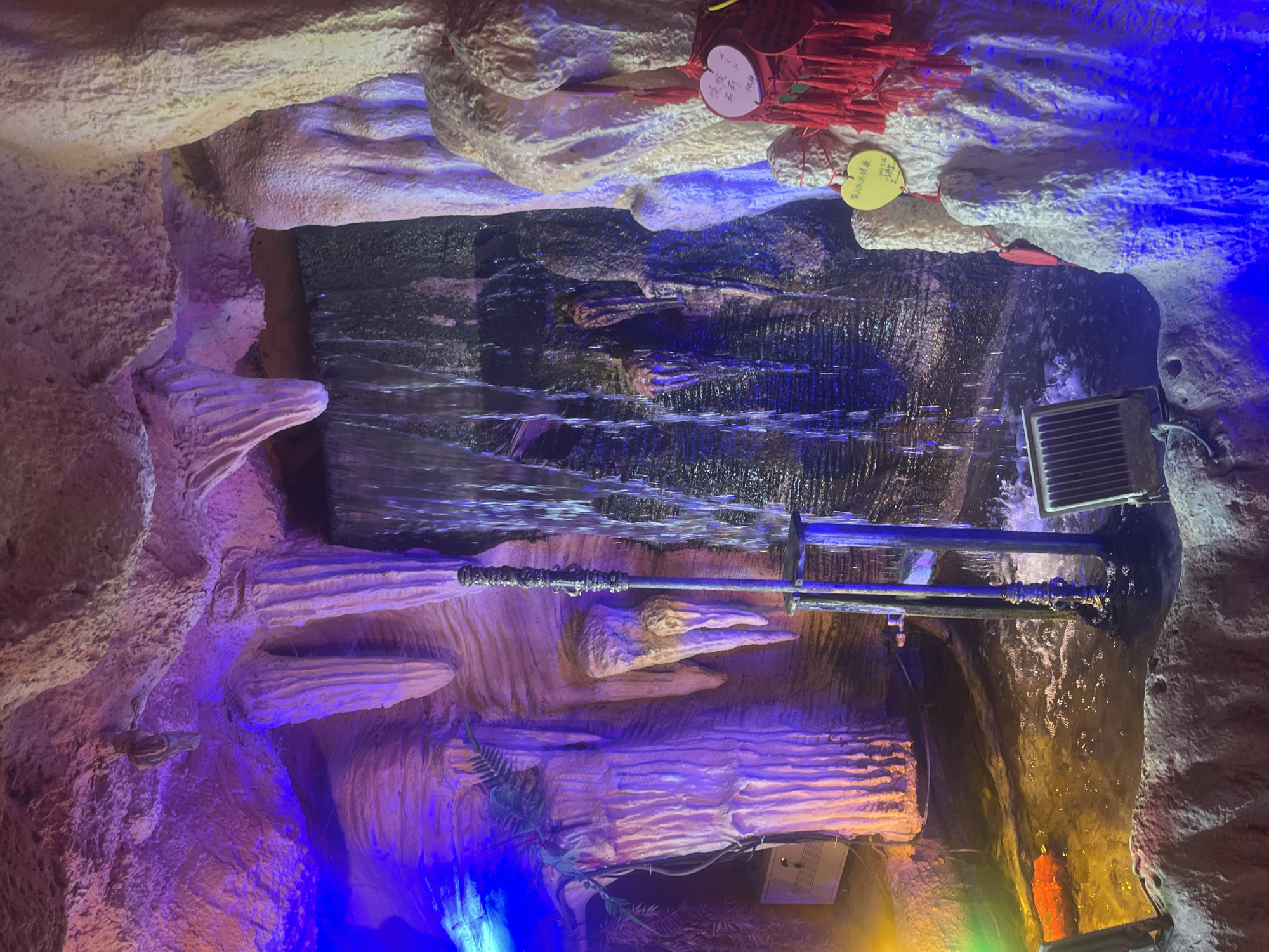 The image depicts an artificial cave or grotto illuminated by vibrant, multi-colored lights, creating a dramatic and fantastical atmosphere. The scene is characterized by rough, textured walls resembling natural rock or stalactite formations, predominantly in shades of beige and gray.
A central feature is a cascading waterfall or flowing water system, created by a series of horizontal structures. Water appears to flow from a higher dark pool, over these structures, and into a lower section. Industrial pipes and metal grates are visible within the water feature, indicating its artificial construction. The water itself reflects the colorful lighting, appearing blue, purple, and green in various sections.
The cave walls are bathed in rich hues: sections are illuminated in deep blue, bright purple, and warm yellow-orange, with some areas showing a subtle green glow. Artificial foliage, resembling ferns or vines, is sparingly placed on some rock ledges, adding to the natural aesthetic. Several spotlights or lighting fixtures are visible, strategically placed to highlight different elements of the display.
Near the top of the image, where the water originates, multiple decorative items are attached to the cave ceiling. These include numerous bundles of red tassels and several heart-shaped tags. Two prominent heart tags are visible: one yellow and one pink. The yellow tag bears Chinese characters that appear to read "平安喜樂" (Píng'ān xǐlè - peace and joy) and "2023". The pink tag also seems to include Chinese characters like "平安" (Píng'ān - peace) and "2023". These tags suggest a cultural practice of writing wishes or prayers.
No people are visible in the image. The setting appears to be an indoor or enclosed tourist attraction, perhaps in Yuzhong District, China, designed to mimic a natural wonder with an added artistic and cultural dimension through lighting and decorations. The time of day is irrelevant as the lighting is entirely artificial.