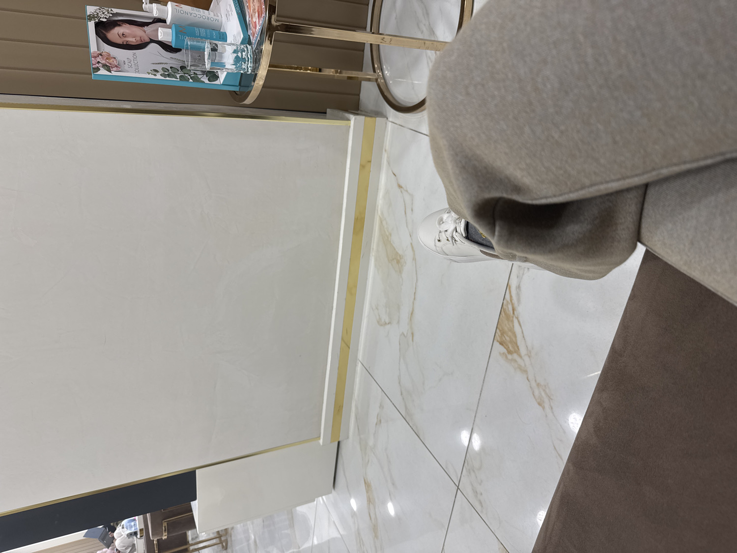 This media file captures an indoor scene from a seated perspective, likely in a modern commercial establishment such as a salon, spa, or upscale waiting area in Sofia, Bulgaria.
In the foreground, a person's legs are visible, covered by light brown or beige trousers. Their left foot, shod in a clean white sneaker with gray socks, rests on a polished white marble-patterned floor. A dark brown upholstered cushion, presumably part of the seat the person is occupying, is partially visible in the bottom right corner.
To the left, a small, round side table with a gold metal frame and a glass top holds several items. On the table, a brochure or magazine cover prominently features a woman's face and the legible text "SCALP COLLECTION" and "MOROCCANOIL." Beside it are multiple small bottles, one clearly labeled "MOROCCANOIL," suggesting hair or beauty products. A clear drinking glass is also present on the table.
The background consists of bright white walls with subtle texture, adorned with horizontal gold trim details. Below these walls, a dark gray or black lower section is visible, possibly a counter or baseboard. The floor, extending throughout the visible area, is made of gleaming white tiles with elegant brown and gold marbling, reflecting overhead artificial light sources.
The overall scene conveys a sense of cleanliness, modern design, and a calm atmosphere, indicating the person is likely resting or waiting for a service in a well-maintained, contemporary space.