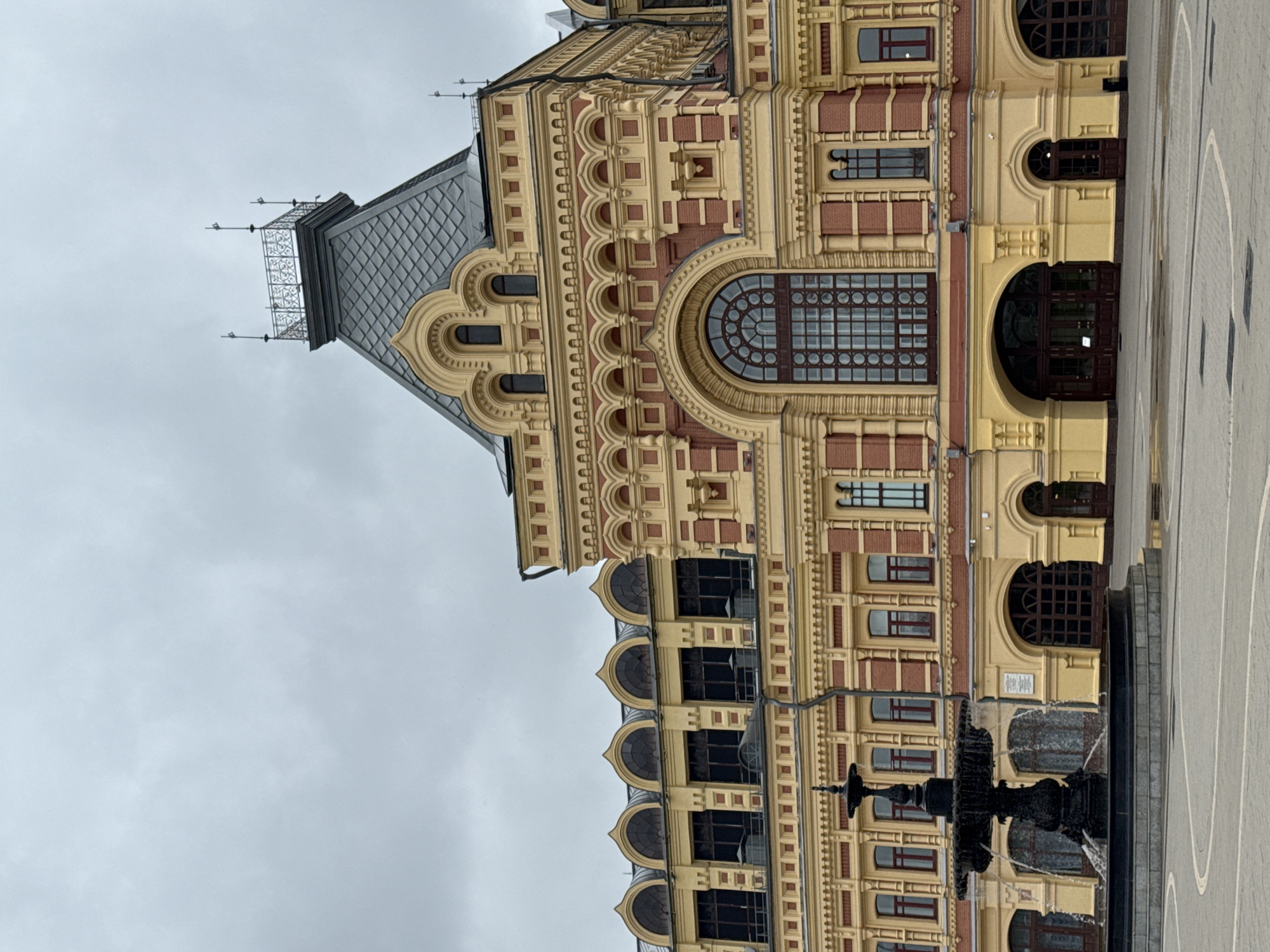 The image depicts a grand, ornate building with a predominantly yellow facade and red brick accents, characteristic of Russian Revival architecture. The building features a prominent central tower with a dark, mansard-style roof, topped by a decorative railing and lightning rods. Several arched windows of varying sizes punctuate the facade, including a large, circular-paned window above the main entrance. The ground level has a series of arched doorways and windows, some with wooden doors.
In the foreground, a dark, multi-tiered fountain with water cascading down is prominently featured. The fountain is set within a circular basin. The ground in front of the building appears to be paved with stone slabs, and there are reflections of water on the ground, suggesting recent rain. The sky is overcast and gray.
This building is the Nizhny Novgorod State Academic Drama Theatre named after Maxim Gorky, located in Nizhny Novgorod, Russia.