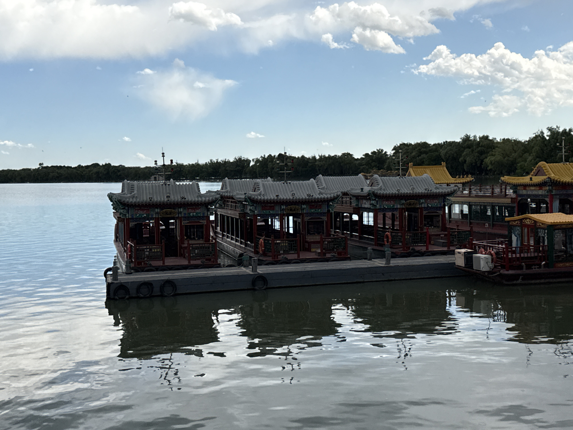 The image displays a tranquil scene on a body of water in Haidian District, China. A pale blue sky, accented with scattered white cumulus clouds, occupies the upper portion of the frame. Below, a distant, flat shoreline is densely covered with dark green trees, forming a continuous band across the midground. The water surface, a muted blue-grey, shows subtle ripples and reflects the sky and the structures along its edge.
In the foreground and midground, a dark grey floating dock, equipped with black cylindrical fenders along its side, extends horizontally. Moored alongside this dock are multiple traditional Chinese-style boats. Several boats feature distinctive grey, tiled, upturned roofs with intricate green, red, and gold decorative elements along their eaves and railings. The main bodies of these boats are predominantly red-brown with visible window openings and railings. Towards the right side of the dock, at least two boats are visible with prominent bright yellow, similarly styled, upturned roofs. Orange life rings are discernable on the railings of some boats. Reflections of the boats and the dock are clearly visible on the water's surface beneath them. No people are present on the boats or the dock.