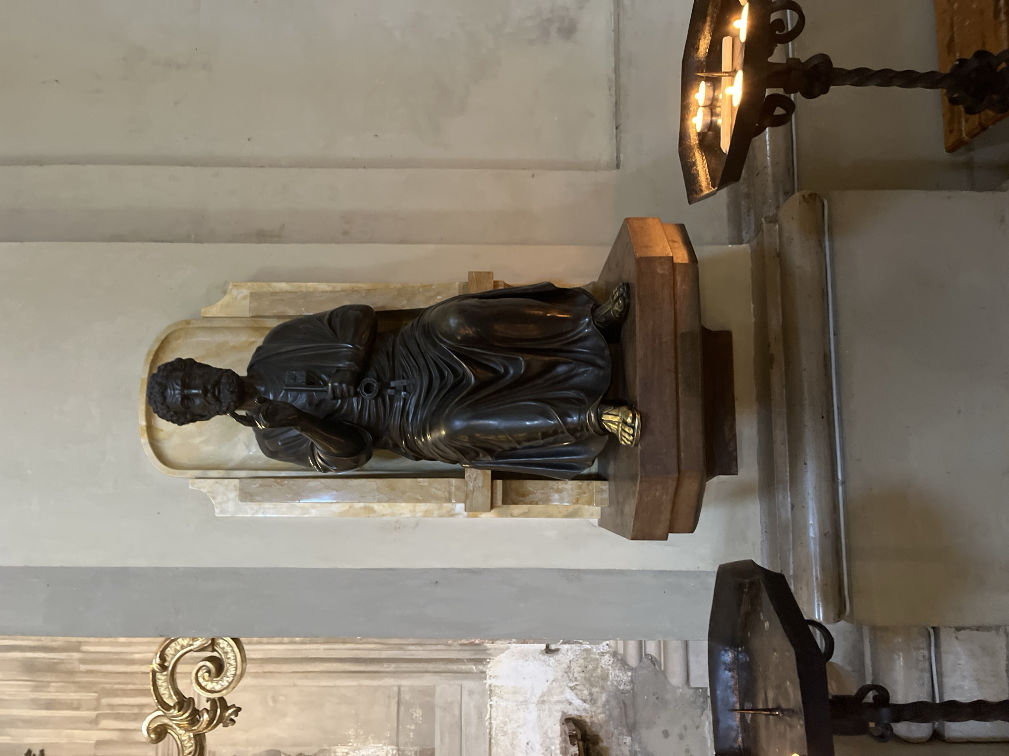A bronze sculpture of Saint Peter with keys rests on a marble base and a wooden stand in a church setting in Bologna, Italy. The sculpture is positioned against a neutral wall with decorative trim. Nearby are ornate metal candle holders. The background includes architectural details of the church, suggesting an indoor, religious environment.