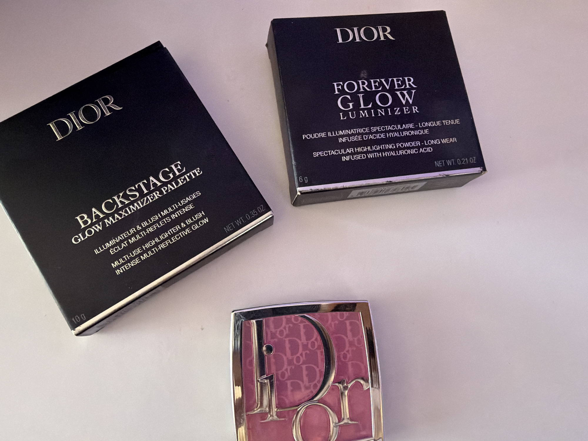 The image shows three cosmetic products from the Dior brand arranged on a white surface. On the left is a black box labeled "DIOR BACKSTAGE GLOW MAXIMIZER PALETTE" with text in both English and French, indicating it's a multi-use highlighter and blush palette. Above and to the right is another black box labeled "DIOR FOREVER GLOW LUMINIZER," also with text in English and French, describing it as a spectacular highlighting powder. Below and in front of the boxes is a pink Dior blush compact with the brand's logo prominently displayed on the surface.