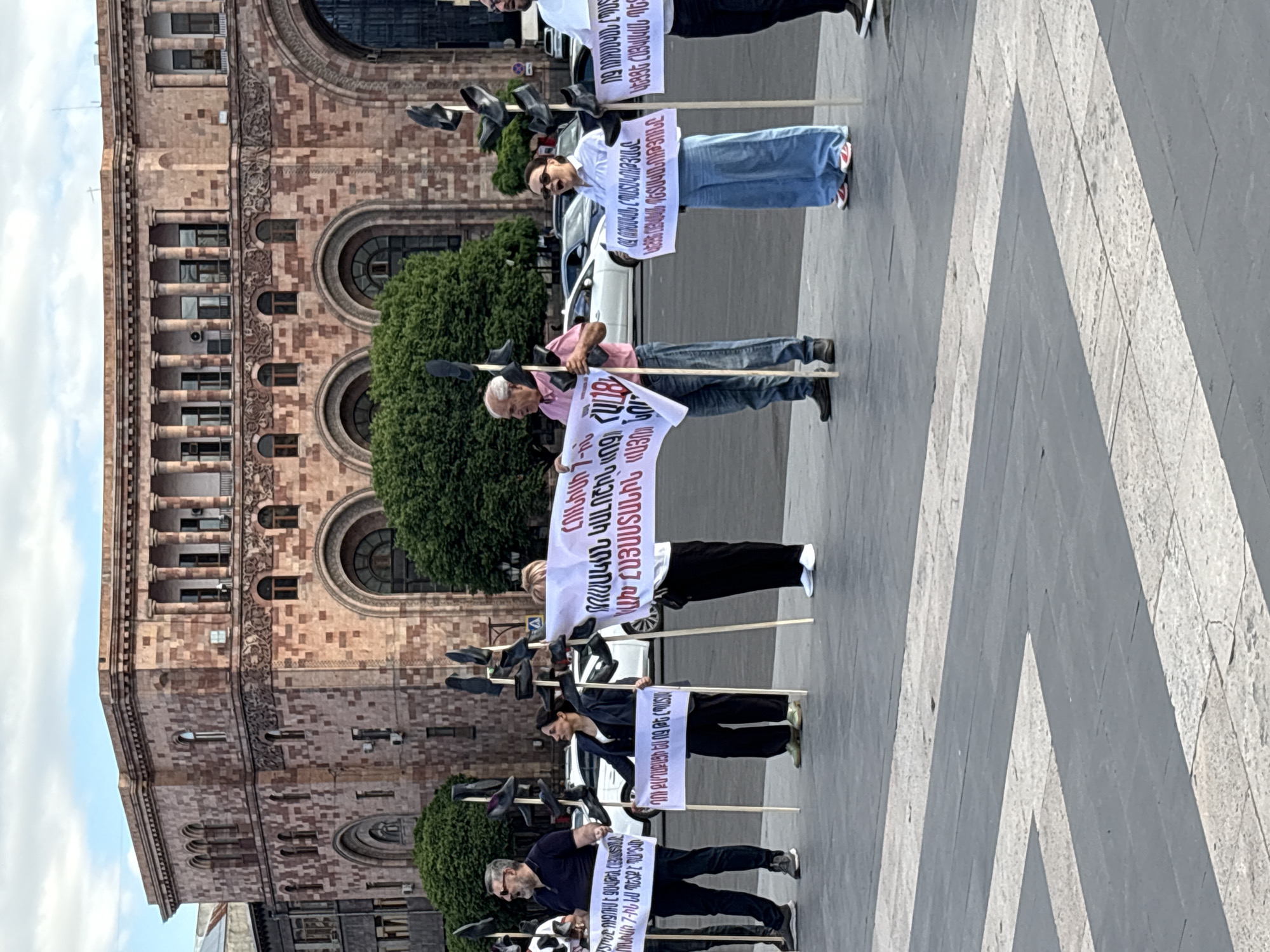 This media file captures an outdoor protest or demonstration taking place during the daytime in Yerevan, Armenia. Several individuals are arranged in a line, holding white banners with text in Armenian. Each person also holds a wooden pole from which multiple black shoes are suspended, a symbolic gesture often used in protests to express strong disapproval or contempt.
The protesters are standing on a paved surface featuring a geometric pattern of grey and light grey tiles. In the background, a large, ornate building with intricate stone architecture is visible. It features multiple stories, columns, arched windows, and decorative carvings, suggesting a significant public or historical building. Green trees partially obscure parts of the building and the background, where white cars are also visible.
The banners display Armenian text, including phrases such as "On June 7, he must lose," "You are a citizen, not a (subject/slave)," and "Long live the Armenian State." Other visible text refers to "Armenia: 'Yes' new." The clear sky with some clouds indicates fair weather during the event. The scene depicts public activism and political expression in a prominent urban setting.