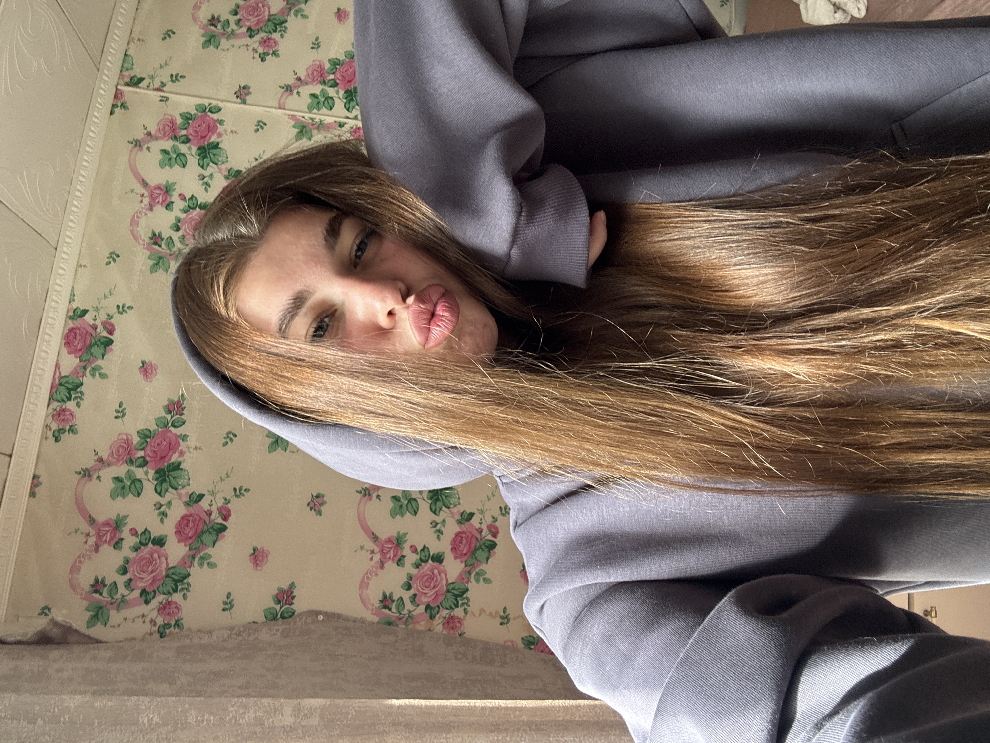 This media file shows a close-up, vertically oriented selfie of a young woman with long, light brown hair. She is wearing a light purple hoodie, with the hood pulled up around her head. Her face is turned towards the camera, and she is making a kissing or puckering expression with her lips. Her eyes are open and looking at the camera, with blue irises. Her eyebrows are dark and well-defined.
The background consists of wallpaper with a floral pattern of pink roses and green leaves on a cream-colored base. There is also a textured white trim running along the top edge of the visible wallpaper. The lighting suggests it is daytime, possibly with sunlight casting shadows, creating a warm glow on her hair and face.
The context provided indicates the location is Мещёры, Russia. There is no visible text in the image. The overall mood conveyed by the selfie is playful and casual.